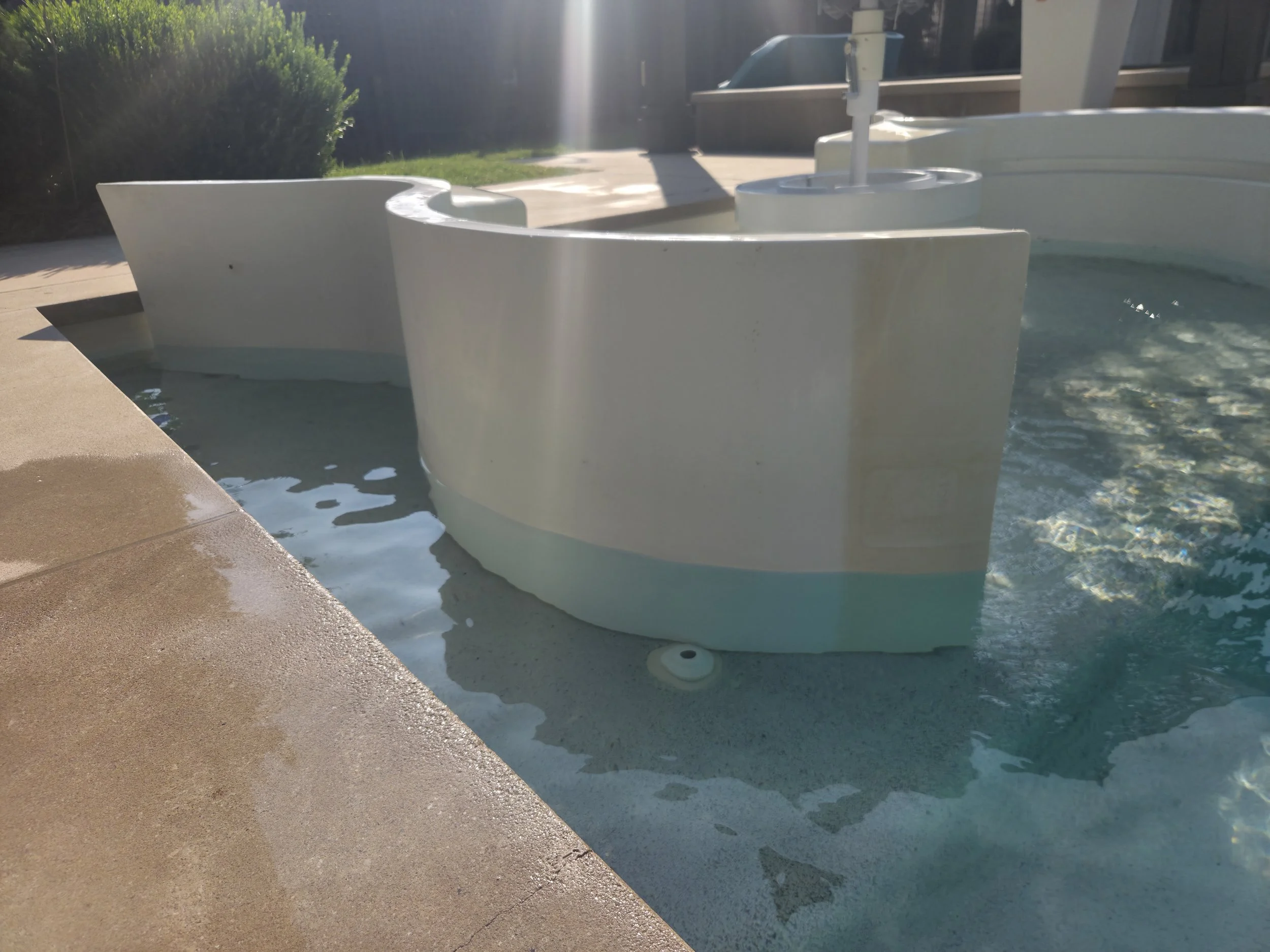 A white hot tub filled with water, located outdoors on a concrete patio with a landscaped yard in the background, sunlight casting shadows.