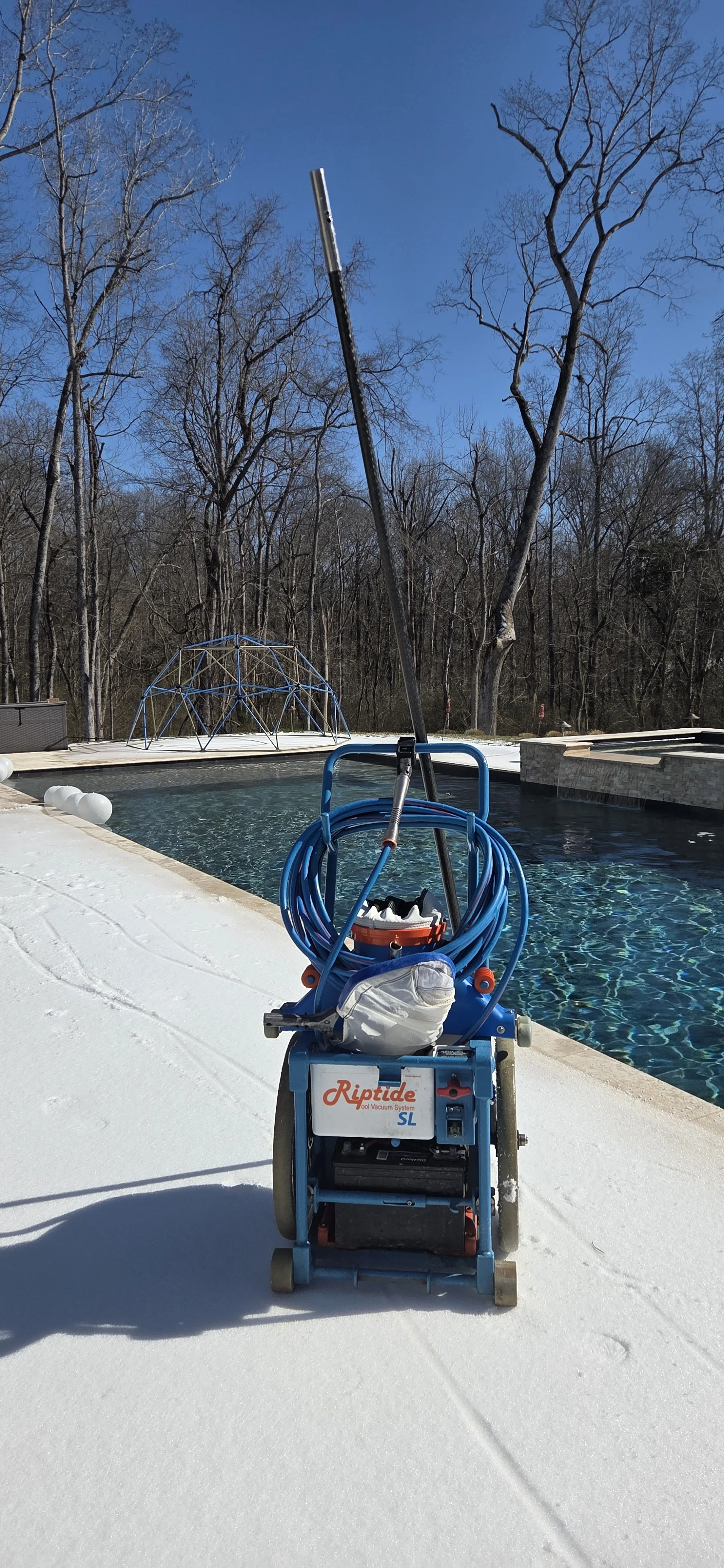 Pool cleaning equipment with blue hose and pole on a snowy deck next to a backyard swimming pool, with a wooded area in the background and a geometric climbing dome at the far end of the pool.