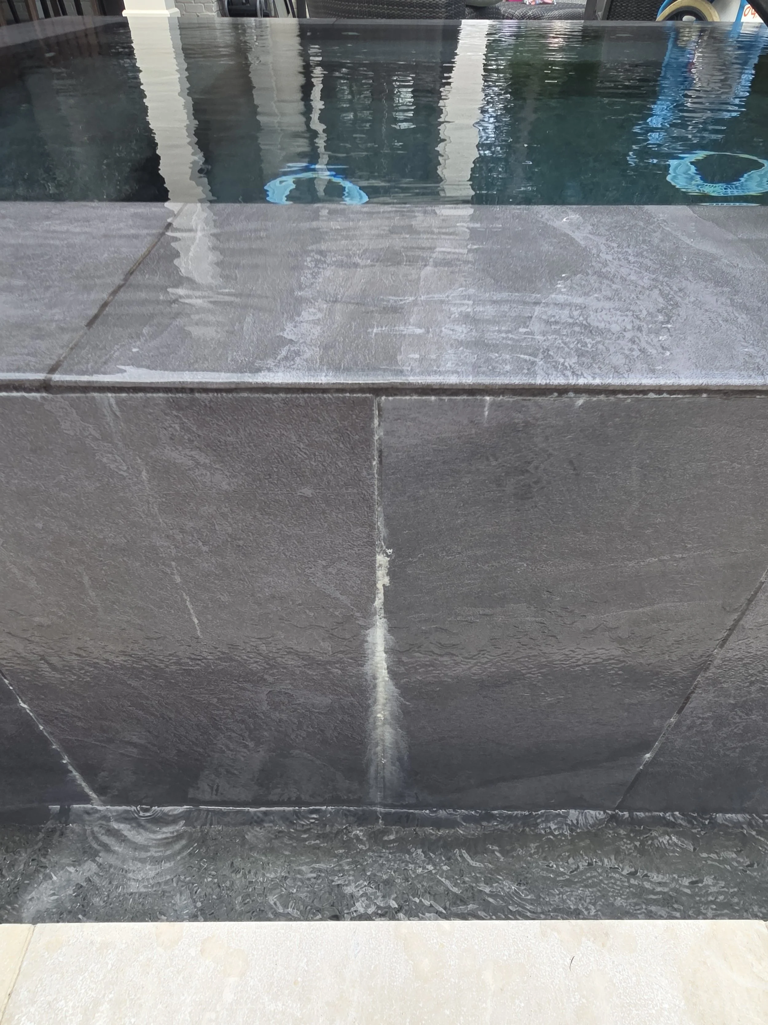 Image of a pool on a patio with a dark stone deck. The pool has clear water reflecting nearby structures, and a small section of beige concrete paving is visible at the bottom.