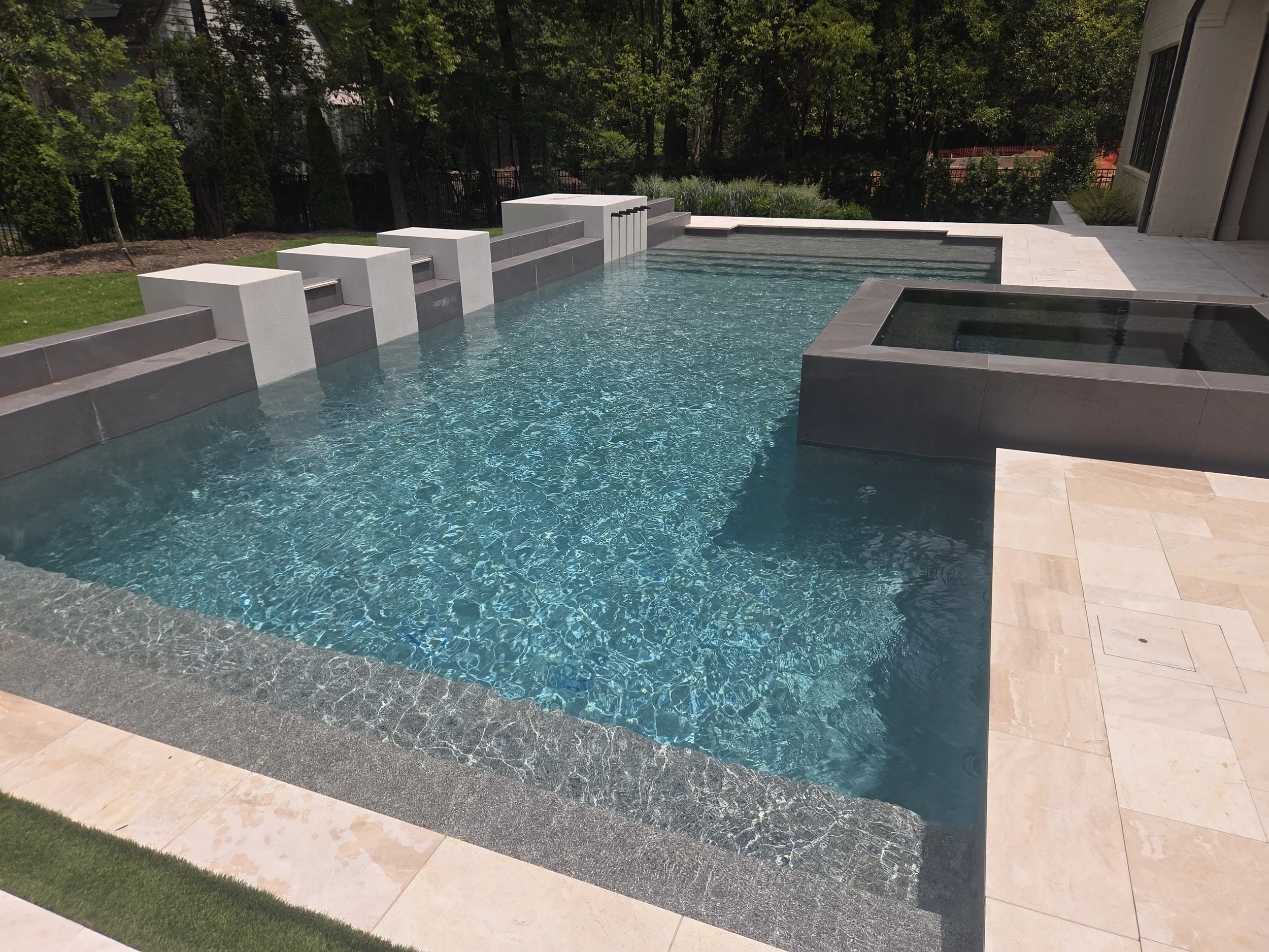 A modern backyard swimming pool with water features, surrounded by greenery and a patio area with light-colored tiles.