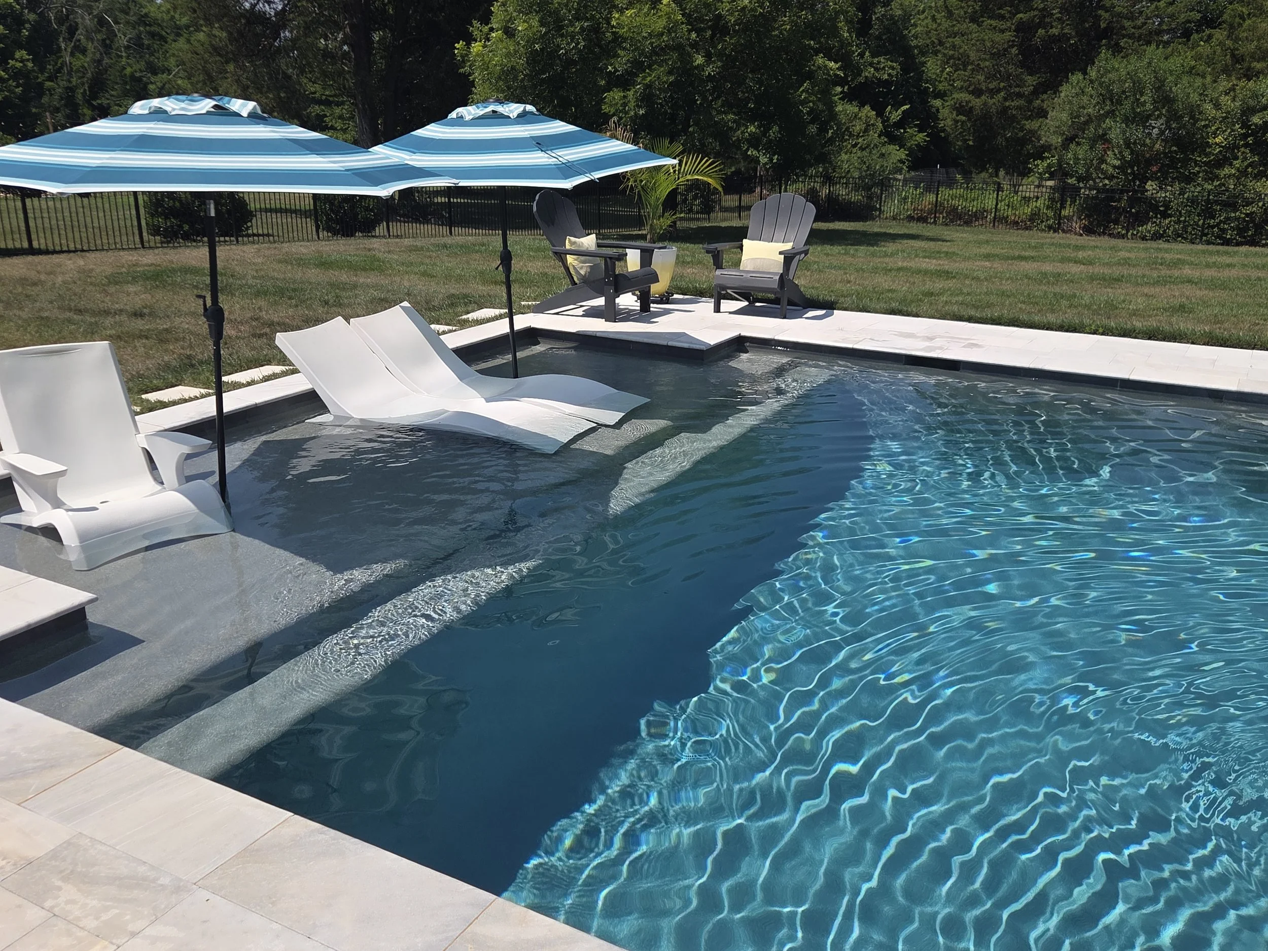 A swimming pool with two white lounge chairs partially submerged, two blue and white striped umbrellas, and two black Adirondack chairs with beige cushions on a concrete deck, surrounded by a grassy yard with trees and a black metal fence.