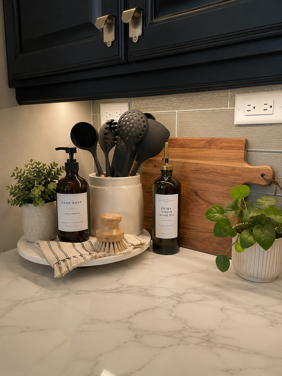 Kitchen countertop with potted plants, hand soap, extra virgin olive oil, a utensil holder with black cooking spoons, and a wooden cutting board.