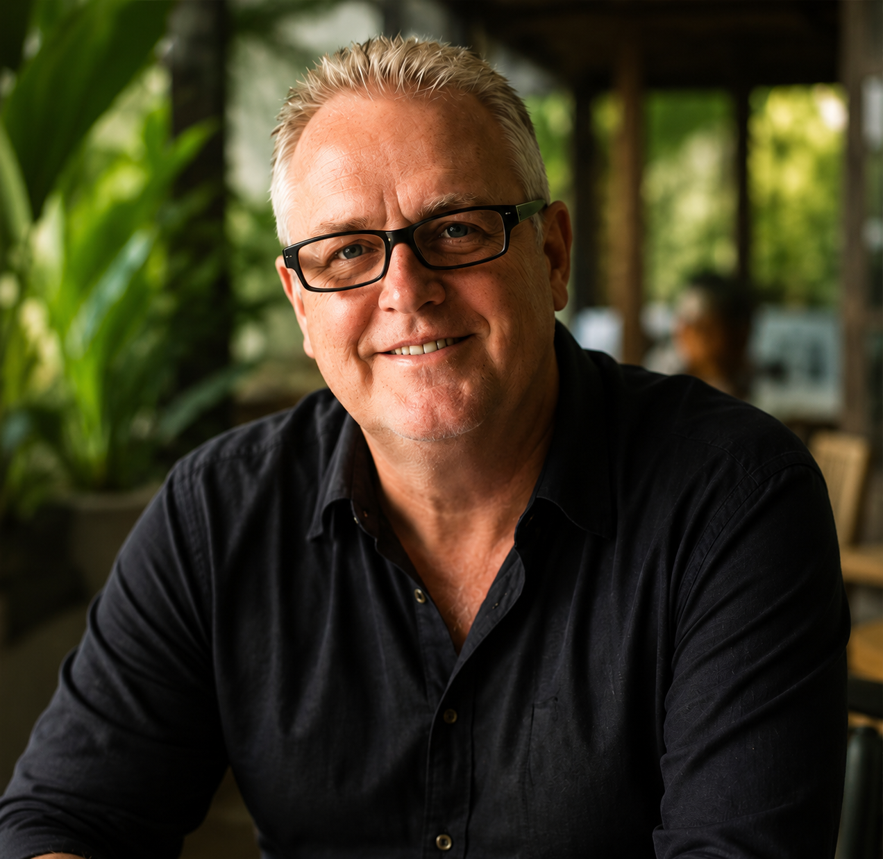 Smiling middle-aged man with glasses, short gray hair, wearing a black shirt, sitting at a table in a green, outdoor restaurant or café.