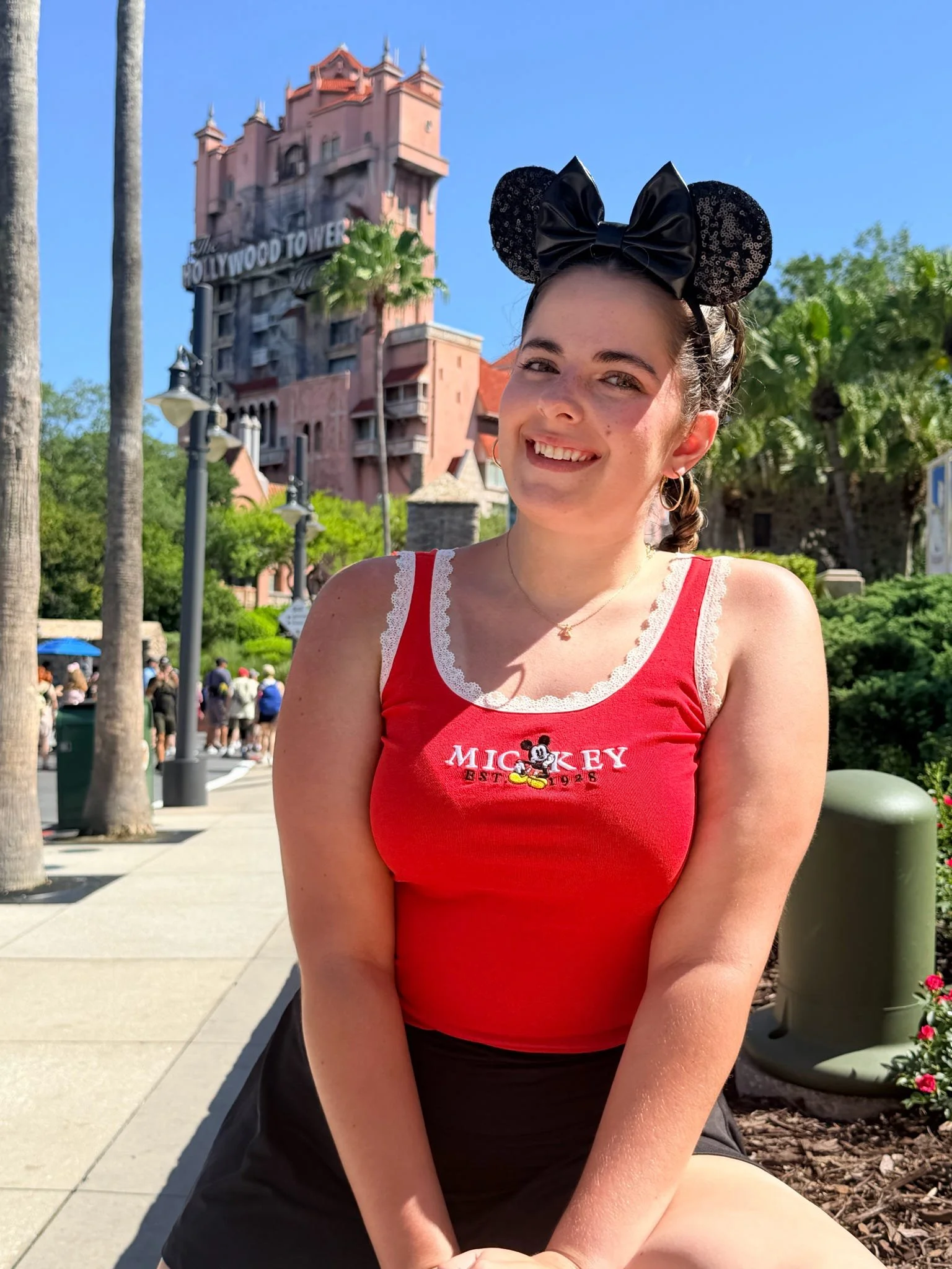 A smiling young woman with a braid, wearing black Mickey Mouse ears and a red tank top, sitting on a sidewalk with lush greenery and a pink castle in the background at a Disney theme park.