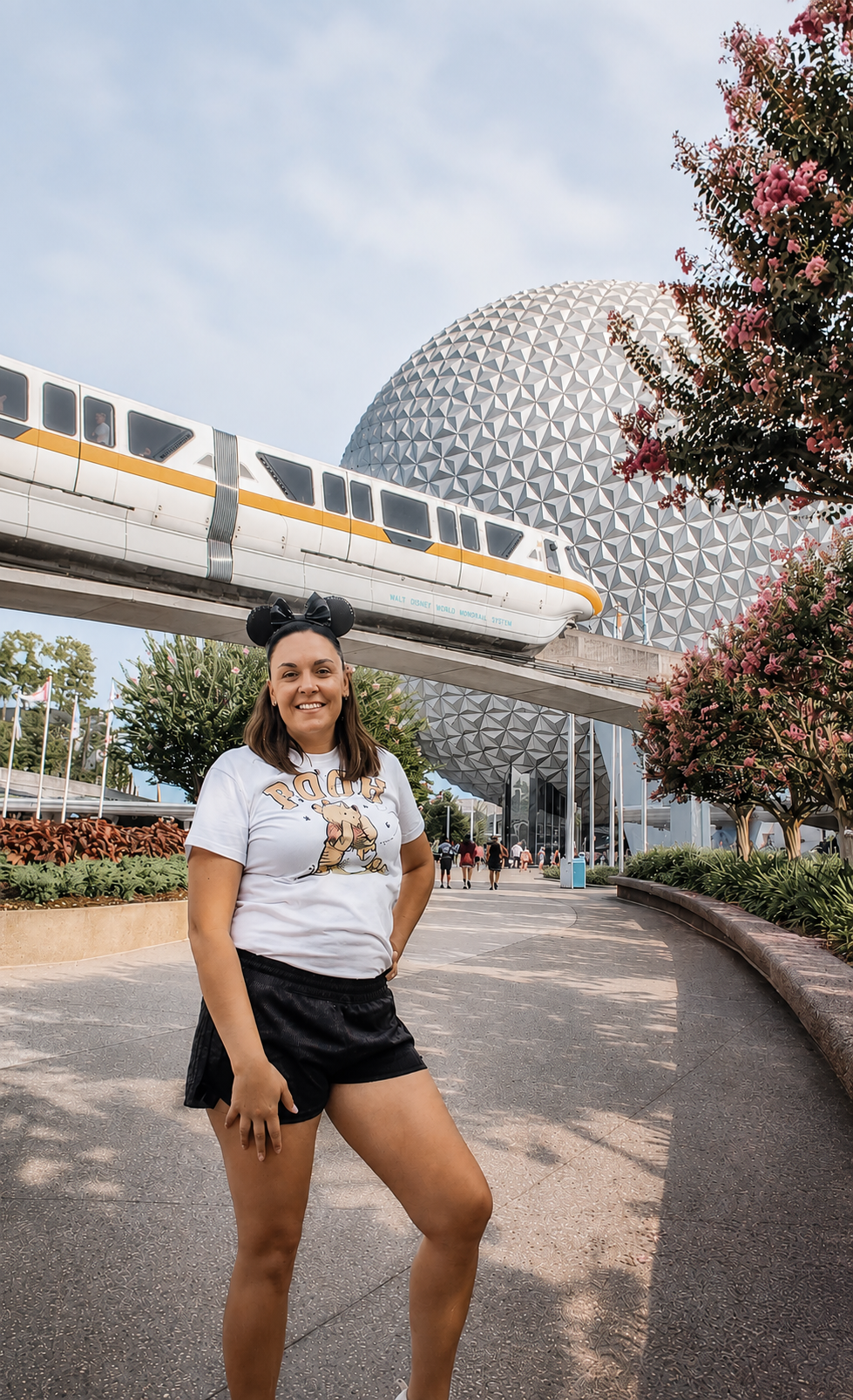 A woman stands in front of Spaceship Earth at EPCOT theme park, wearing a white t-shirt with a cartoon character, black shorts, and Mickey Mouse ears hat, smiling at the camera.