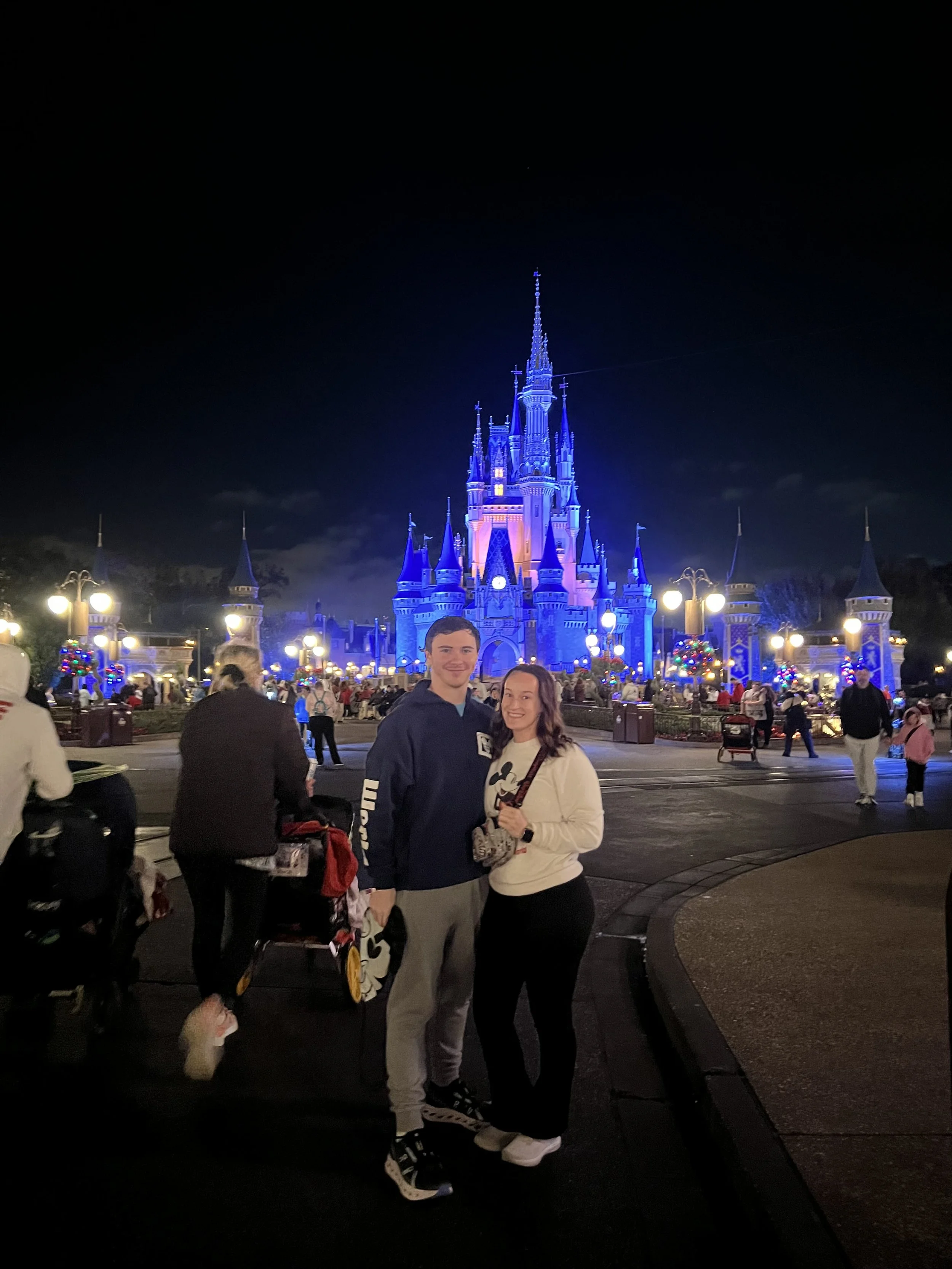 A young couple stands in front of Cinderella's Castle at night, illuminated in blue lights, at a Disney theme park. The area is bustling with visitors.