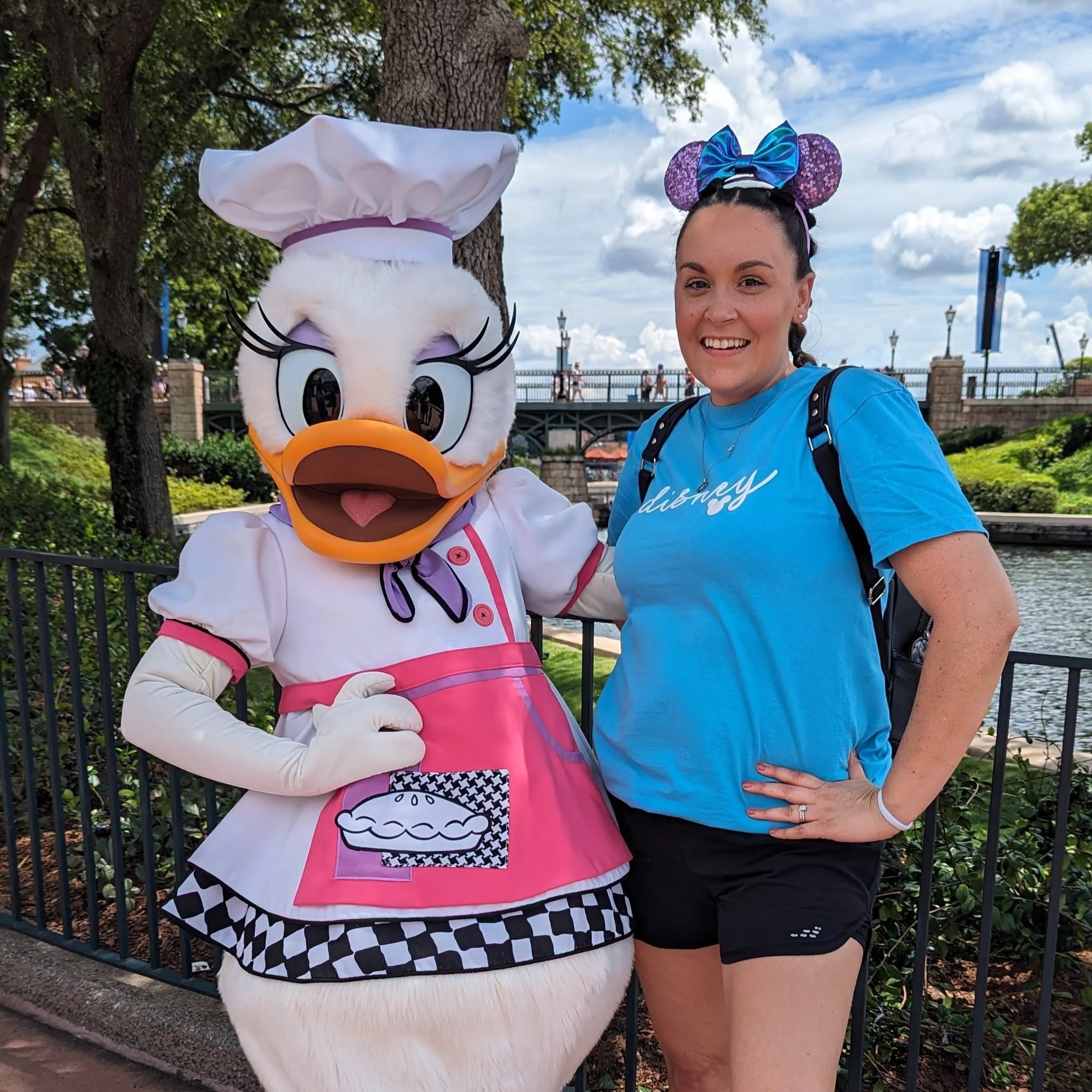Daisy Duck taking a selfie with Kim Norton wearing a blue Disney shirt and Minnie Mouse ears headband outdoors near a river, trees, and a bridge.