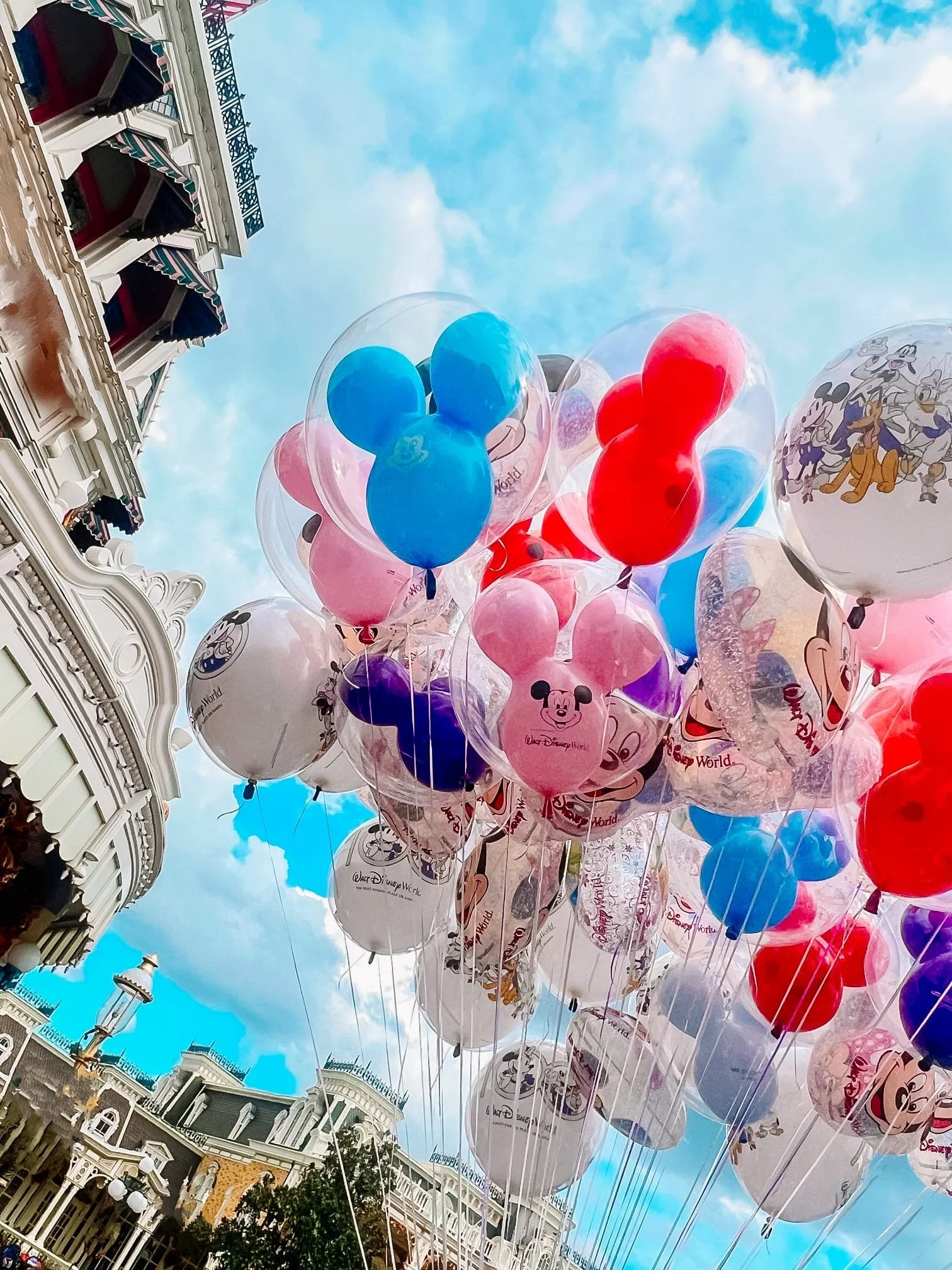Colorful balloons featuring Mickey Mouse and Disney characters floating in the sky above a city street lined with Victorian-style buildings on a partly cloudy day.