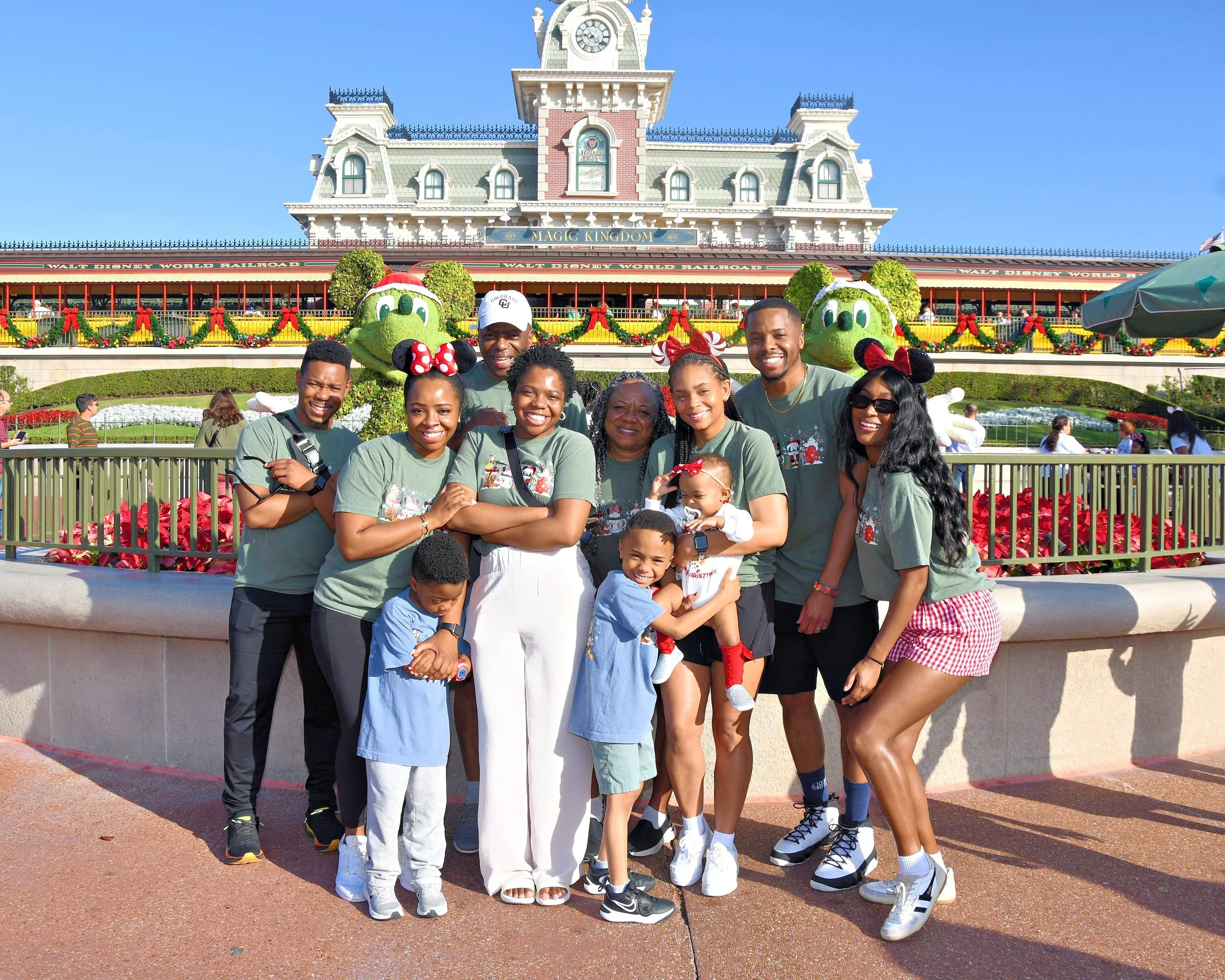 A diverse group of ten people, including children and adults, standing in front of the Disneyland entrance with a large clock tower and holiday decorations, smiling and posing for a photo.