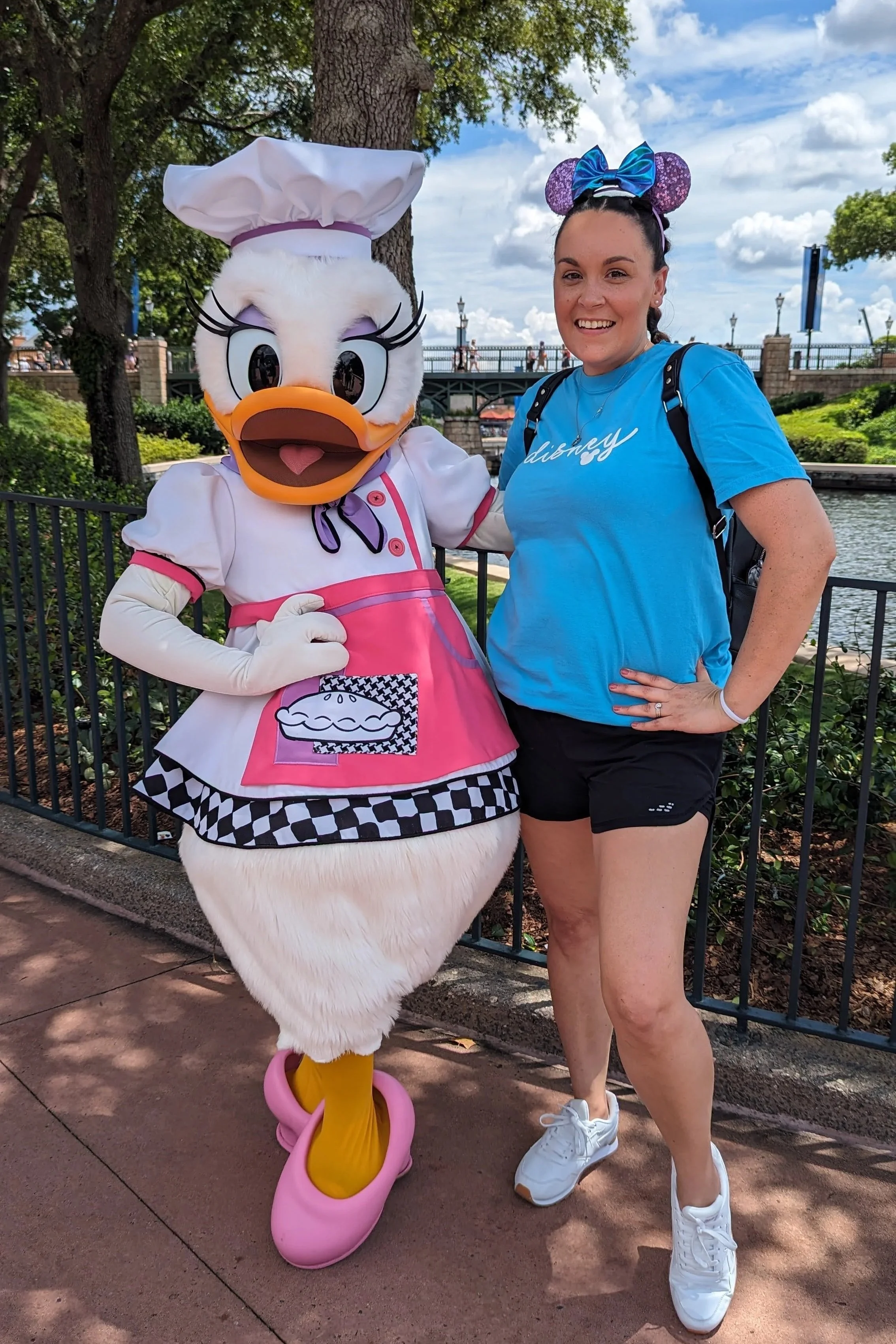 Kim Nortondressed in casual Disney-themed attire posing next to a costumed character of Daisy Duck dressed as a chef at a Disney park.