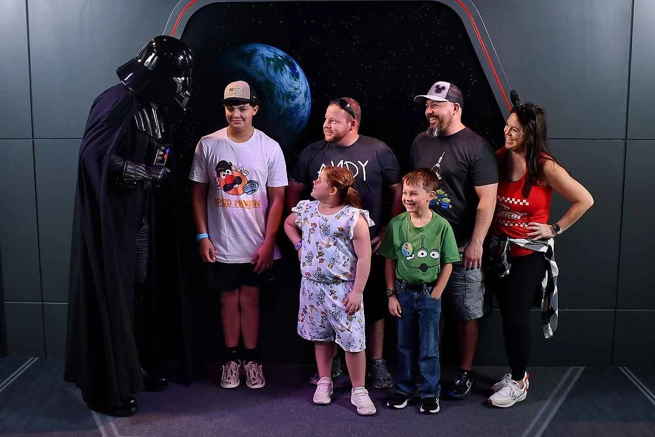 People taking a photo with Darth Vader at a themed exhibit. The group includes three children and three adults, with a space-themed background showing Earth and outer space.