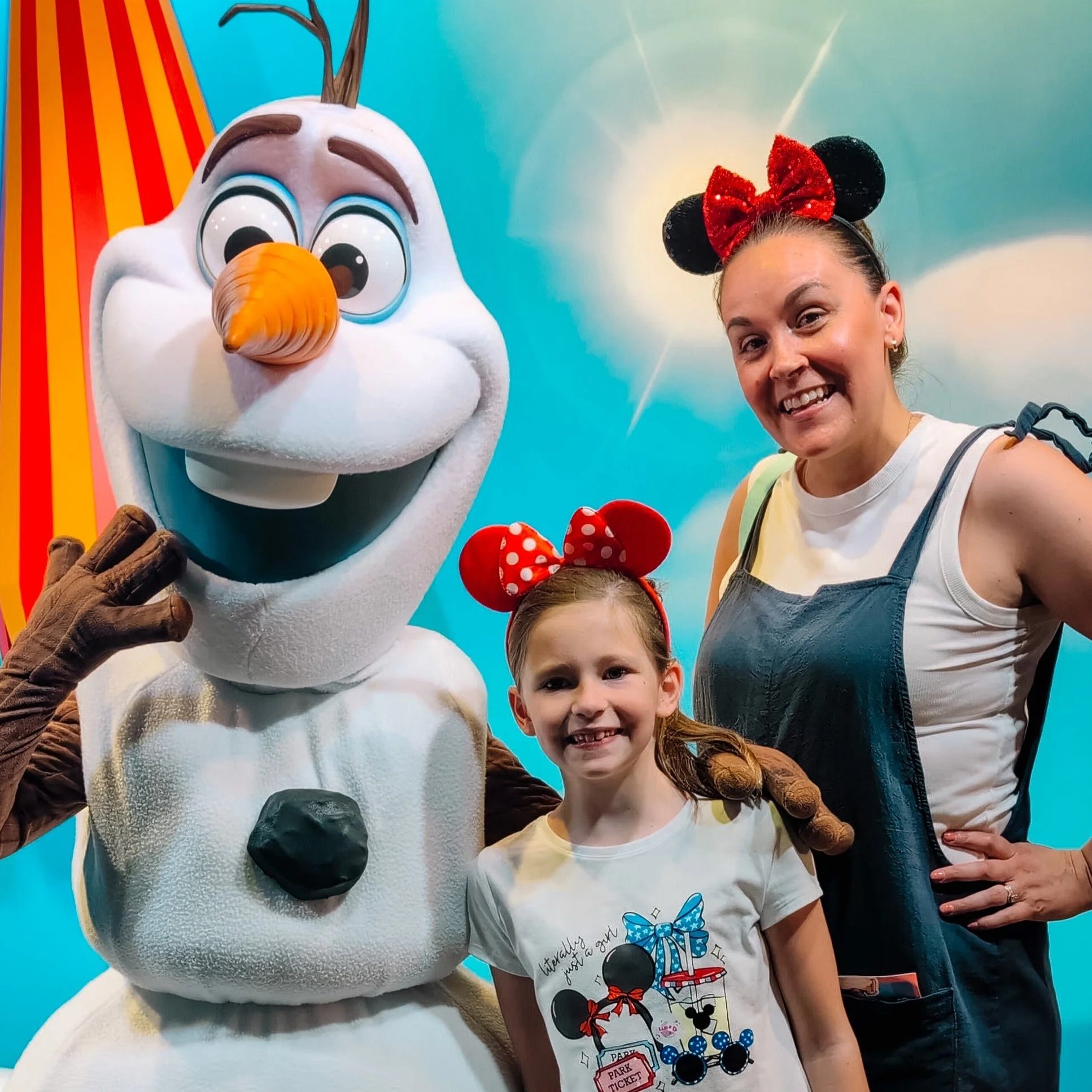 A woman and two children posing with Disney characters Olaf and Minnie Mouse at a theme park. Olaf is a snowman with a large orange carrot nose and twig hair, and Minnie Mouse is wearing a red polka-dotted bow and dress. The background features a blue sky with a subtle star motif.