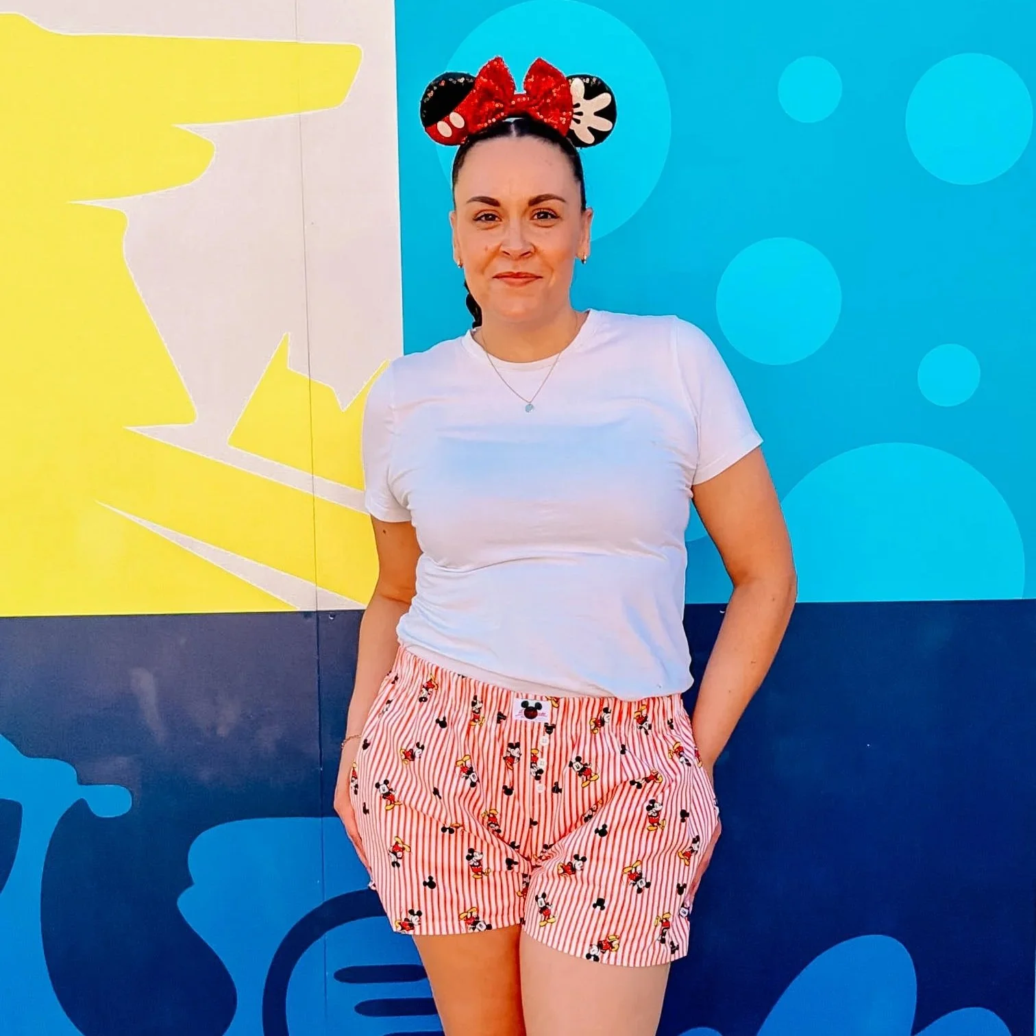 A woman standing in front of a colorful wall with blue and yellow sections, wearing a white T-shirt, Disney Mickey Mouse patterned shorts, and a Mickey Mouse ears headband with a red bow.