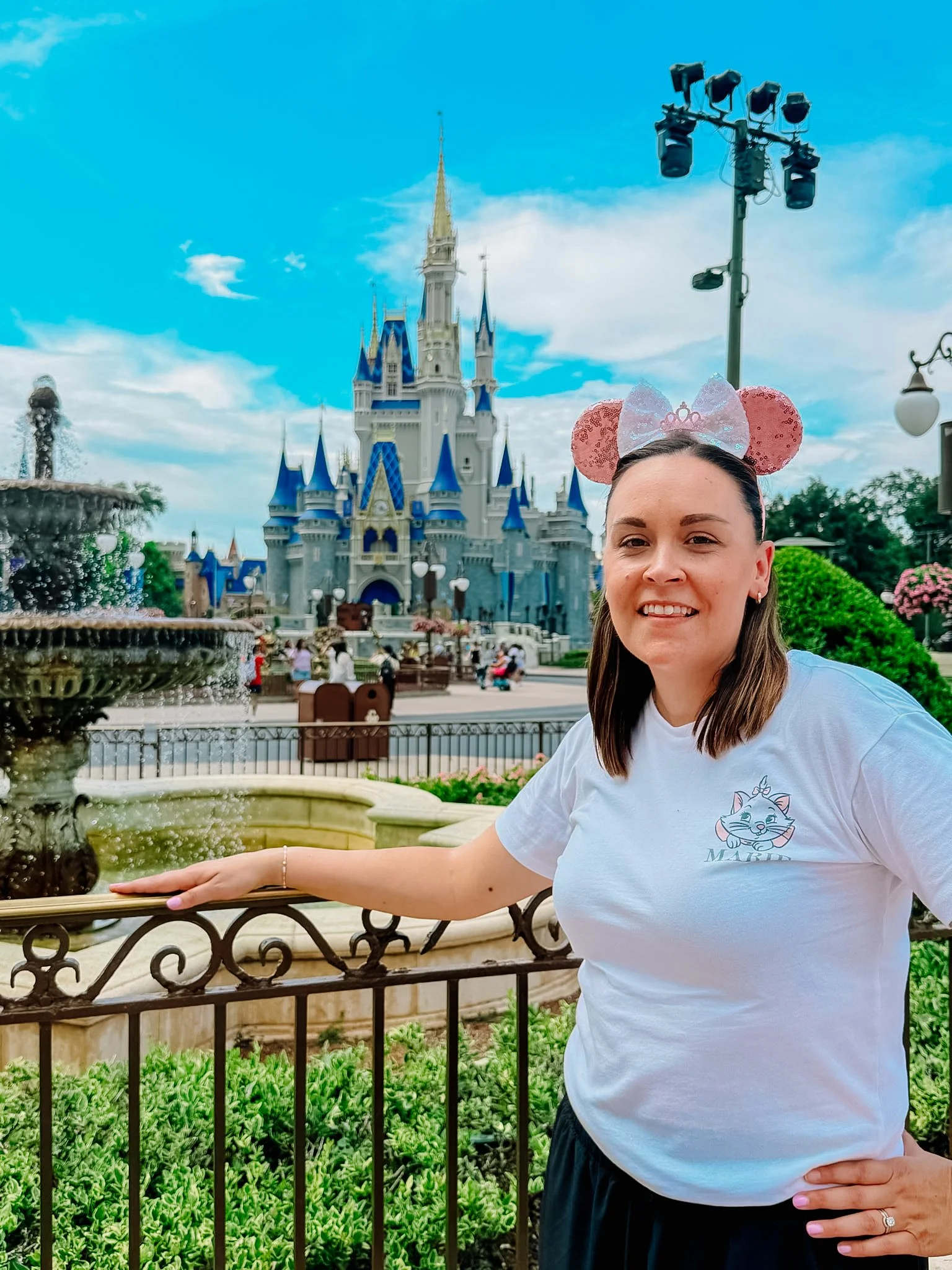 A woman wearing Minnie Mouse ears headband, smiling, standing near a fountain at a Disney theme park with Cinderella's castle in the background.
