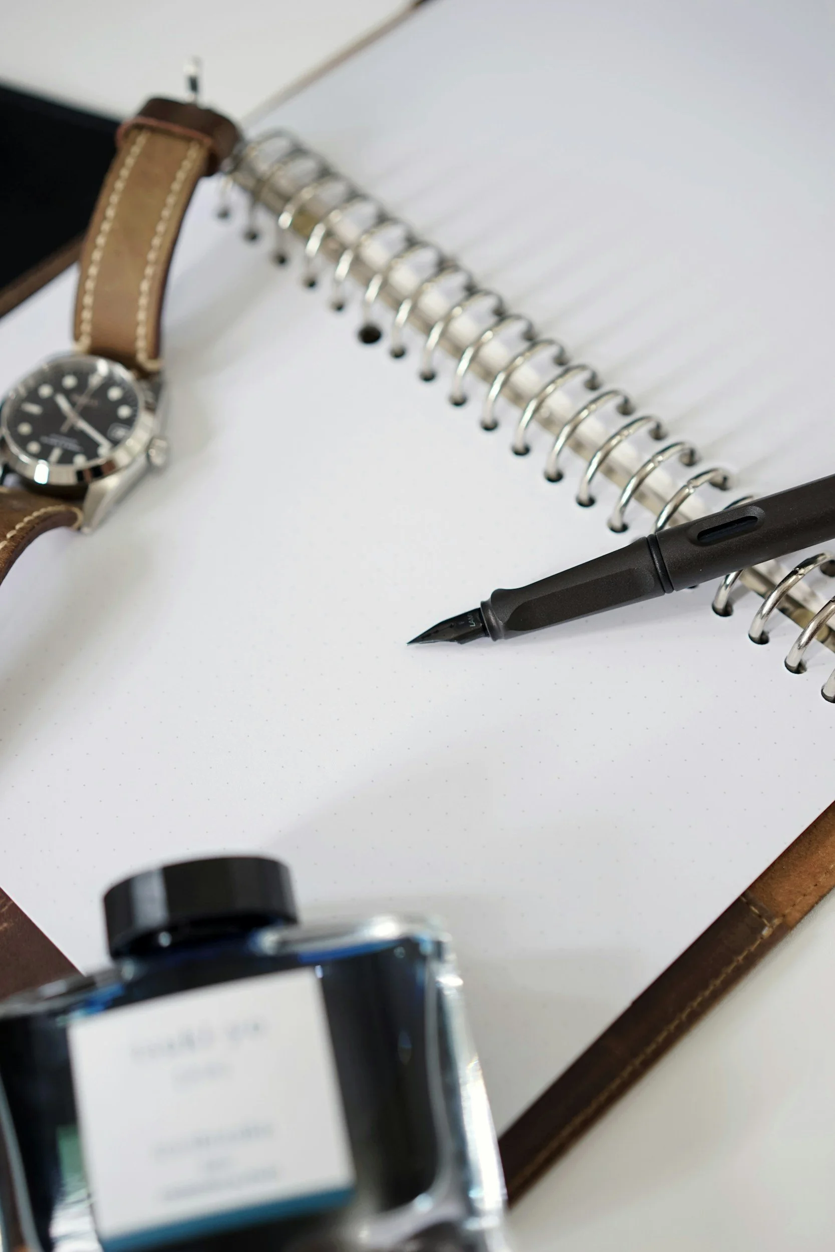 Close-up of a black pen on a spiral notebook with a wristwatch and other documents on a leather surface.