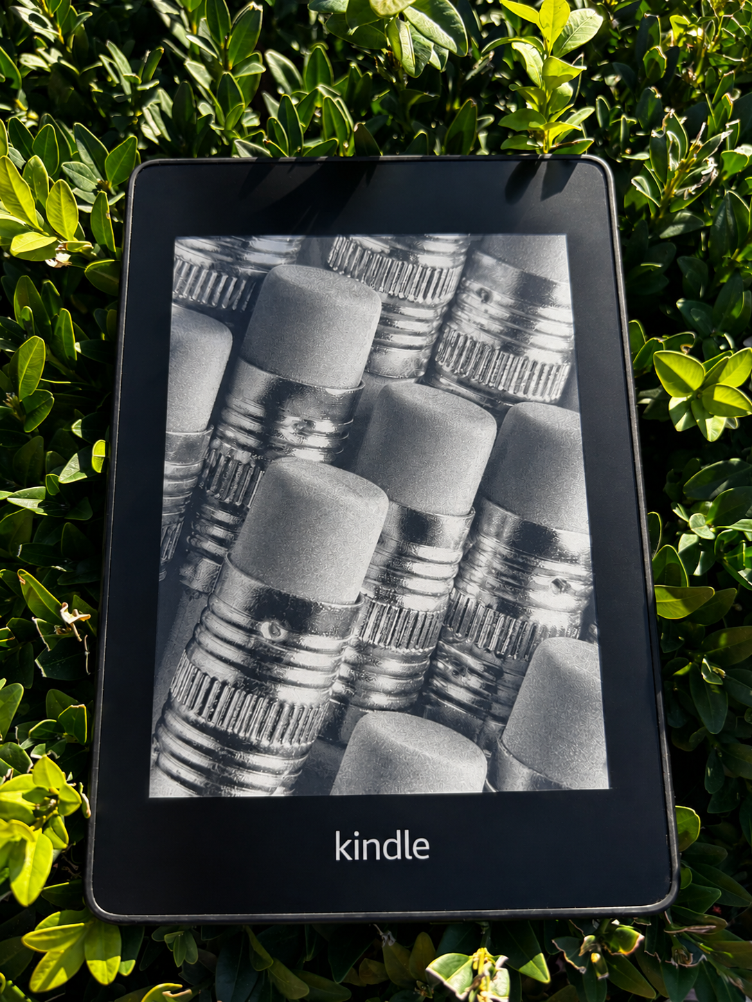 An Amazon Kindle e-reader placed on green bushes, displaying a black and white image of pencils.