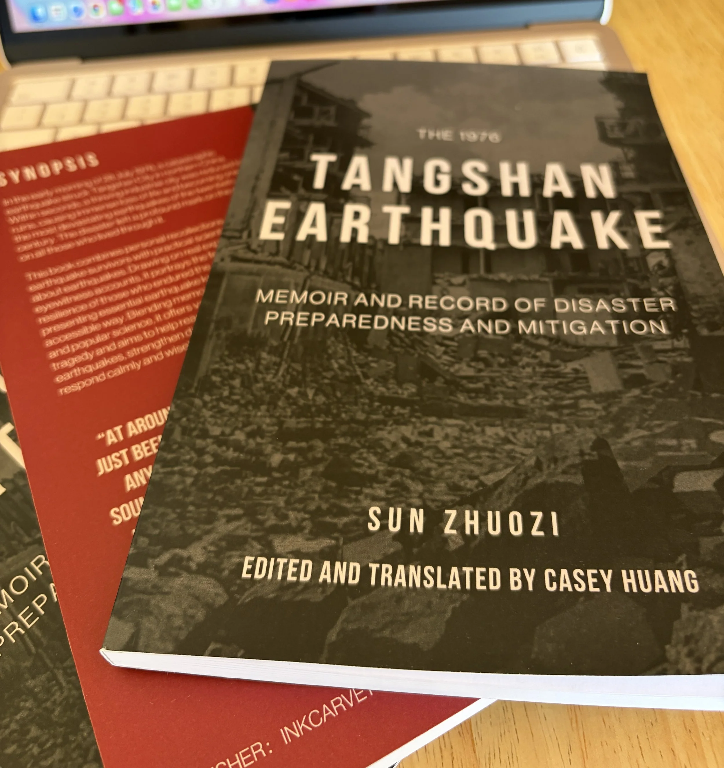 Printed edition of The 1976 Tangshan Earthquake: Memoir and Record of Disaster Preparedness and Mitigation displayed on a work desk with a laptop in the background.