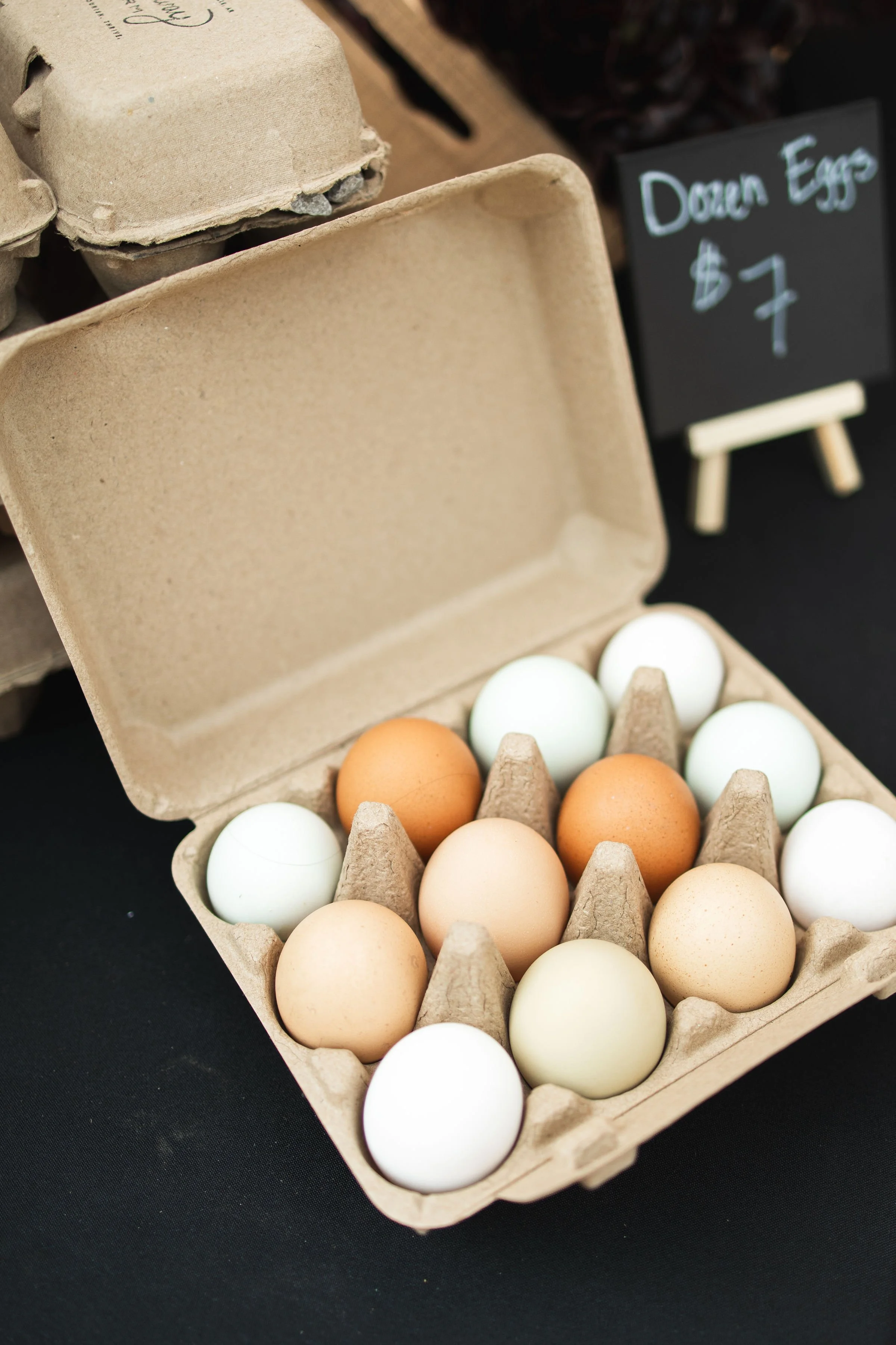 A carton of mixed brown, white, and pale green eggs is open on a black surface at a market stall, with a small chalkboard sign in the background indicating that the eggs are priced at $7.