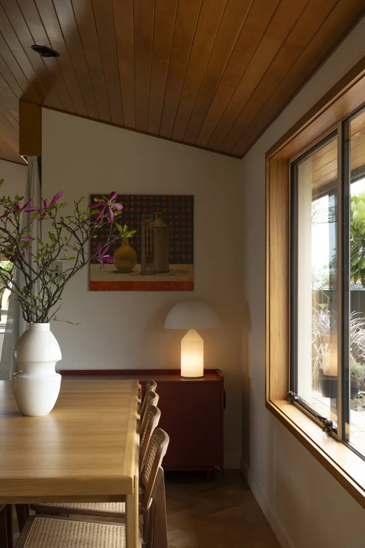 Interior view of a dining room featuring a wooden table with chairs, a white vase with pink flowers, a sideboard with a modern lamp, a framed artwork on the wall, and a window with a wooden frame showing outdoor greenery.
