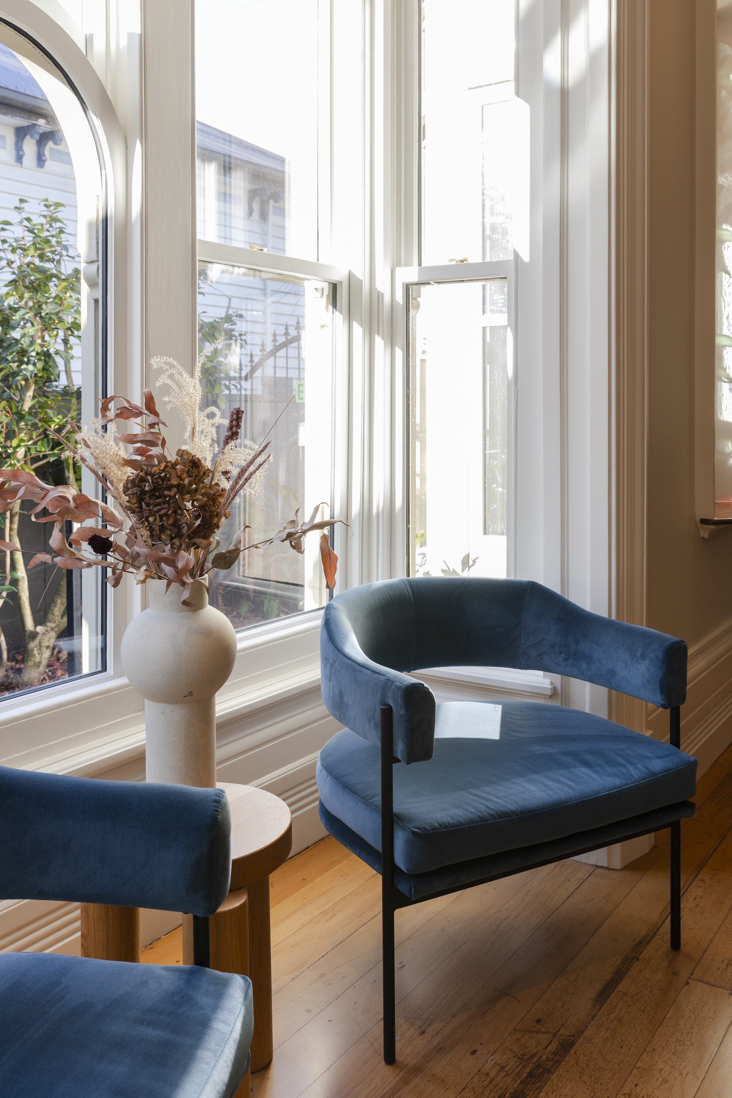 A cozy corner of a room with wooden floors and sunlight streaming through large windows. There are blue velvet chairs and a white vase with dried flowers on a small round table.