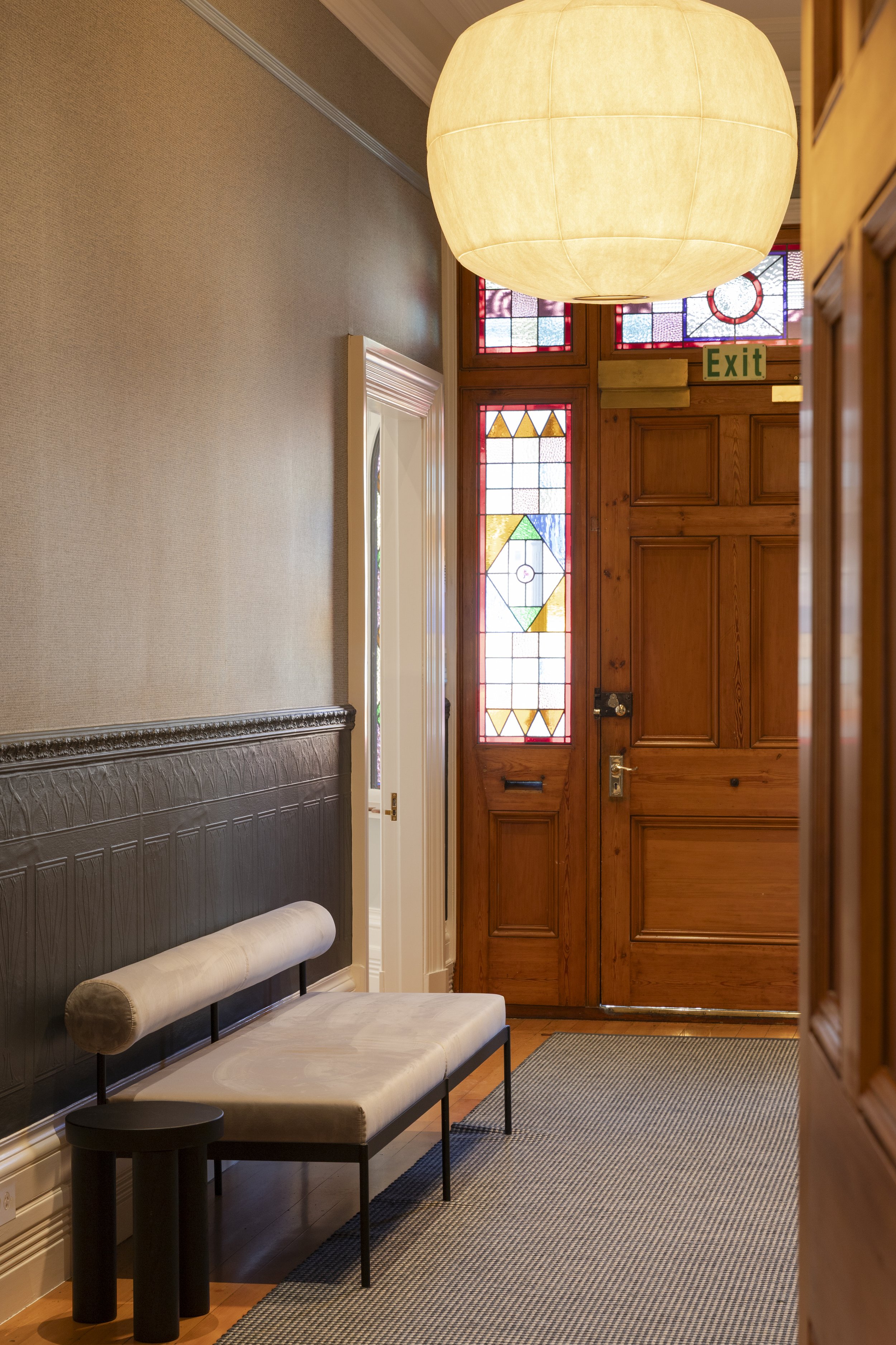 Interior of a room with a wooden door, stained glass windows, a beige bench with a cylindrical cushion, a small round black side table, and a large round pendant light hanging from the ceiling.