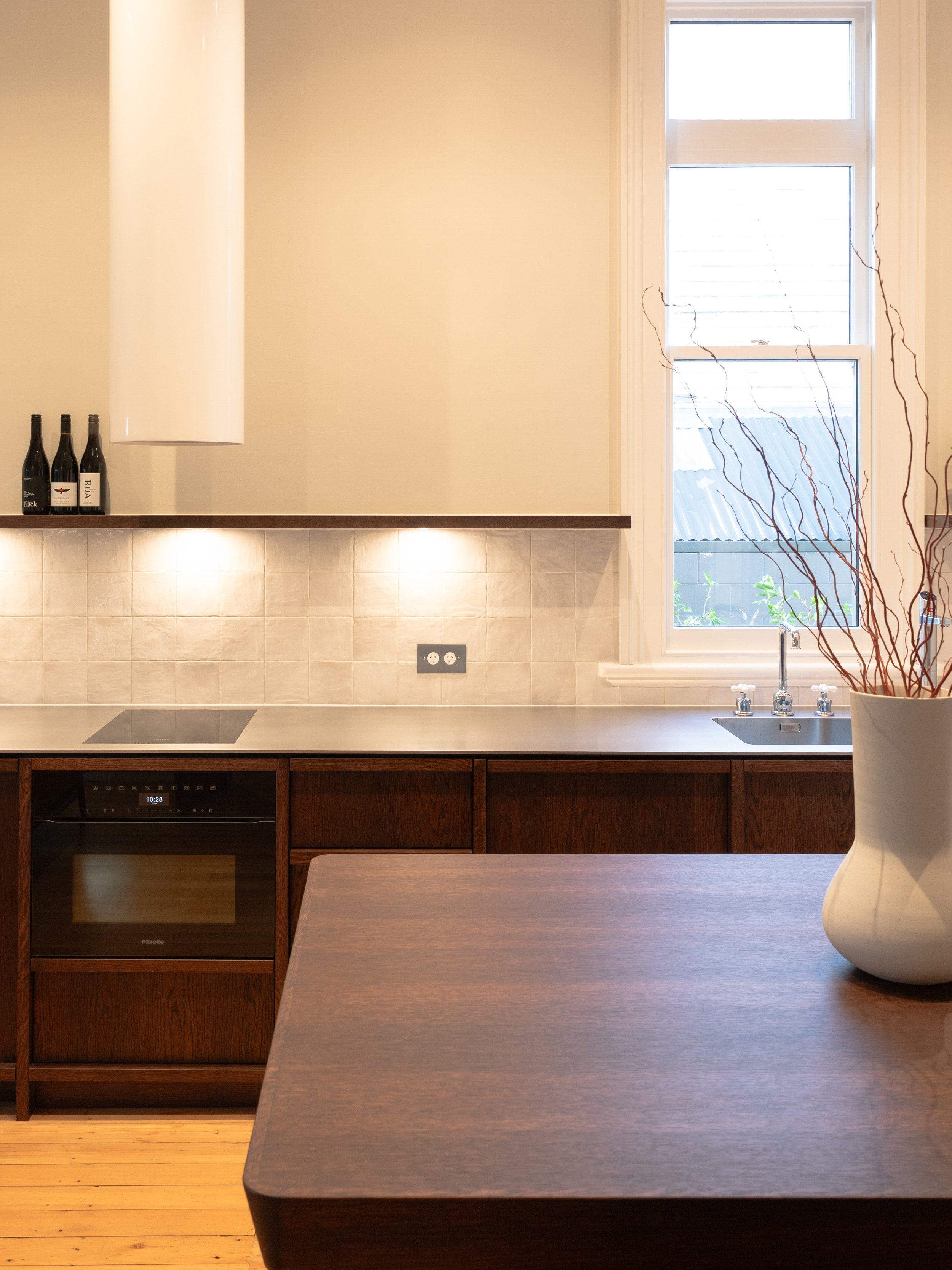 A kitchen with a window, a white vase with twigs, bottles of wine, a black oven, and a wooden countertop.