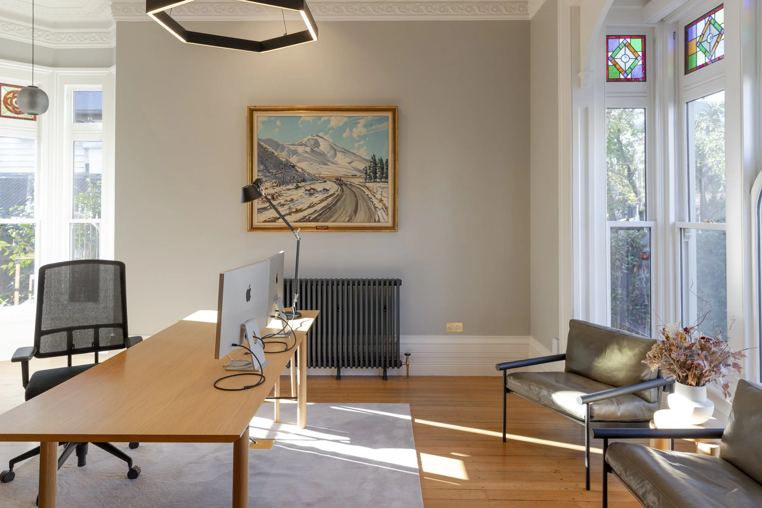 Office space with a desk containing an Apple iMac, a black mesh office chair, and a floor lamp. The room has large windows with stained glass accents and a framed painting of a snow-covered mountain landscape on the wall. There is a black radiator and a beige armchair with a side table holding a vase of dried flowers.