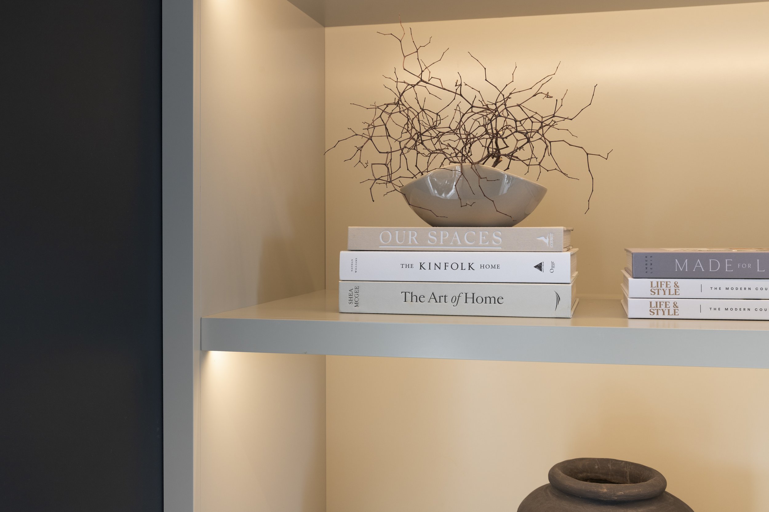 A beige-colored shelf displays several coffee table books stacked horizontally with titles such as "Our Spaces," "The Kinfolk Home," and "The Art of Home." On top of the books, there's a ceramic vase holding a dried, leafless branch arrangement. The background wall is painted a neutral tone, creating a minimalist aesthetic.