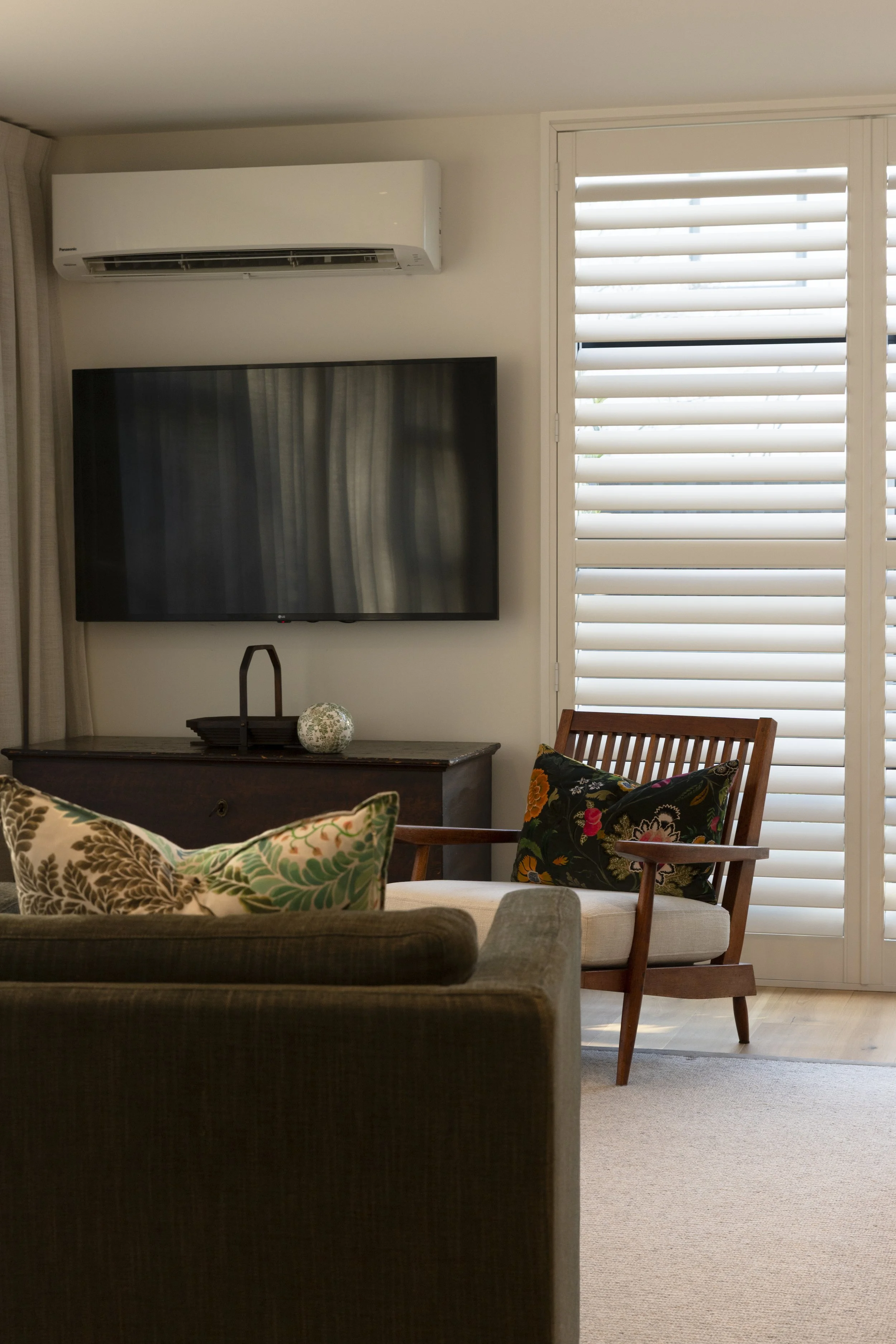 Living room with a wall-mounted flat-screen TV, an air conditioning unit above, a wooden armchair with floral pillows, a wooden side table with a decorative ball, and a window with white horizontal blinds.