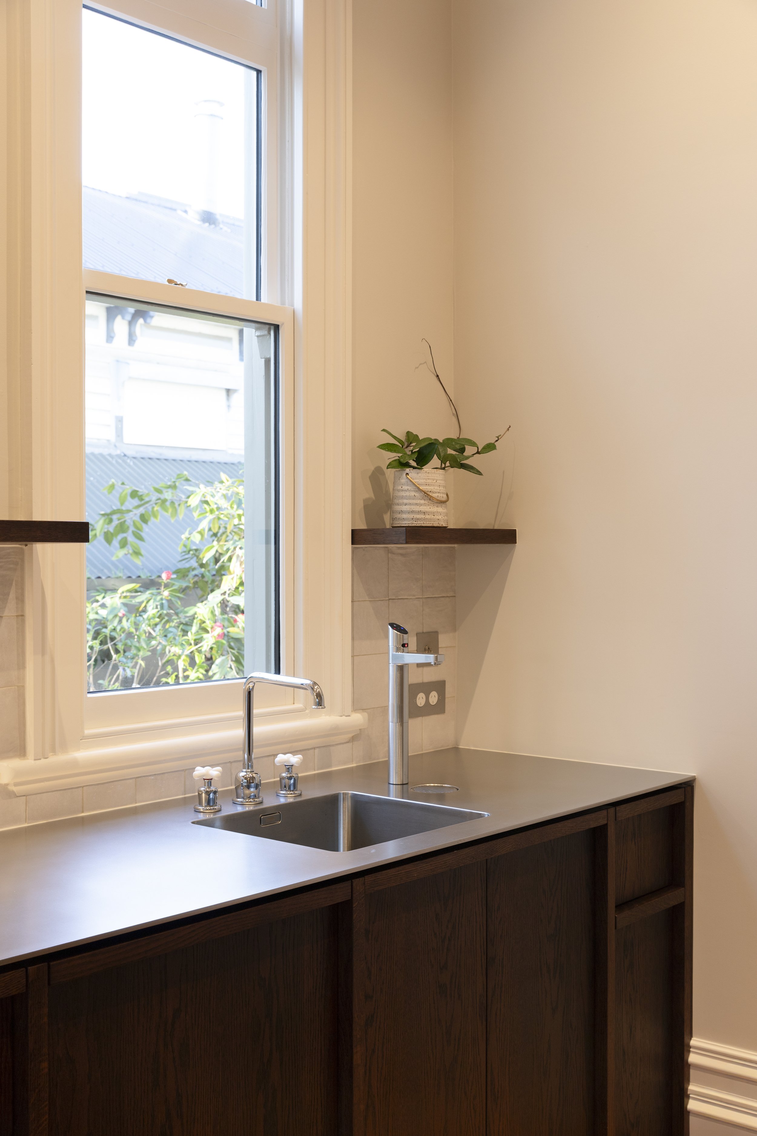 Kitchen sink with a view of a window, a potted plant on a floating shelf, and dark wooden cabinetry.