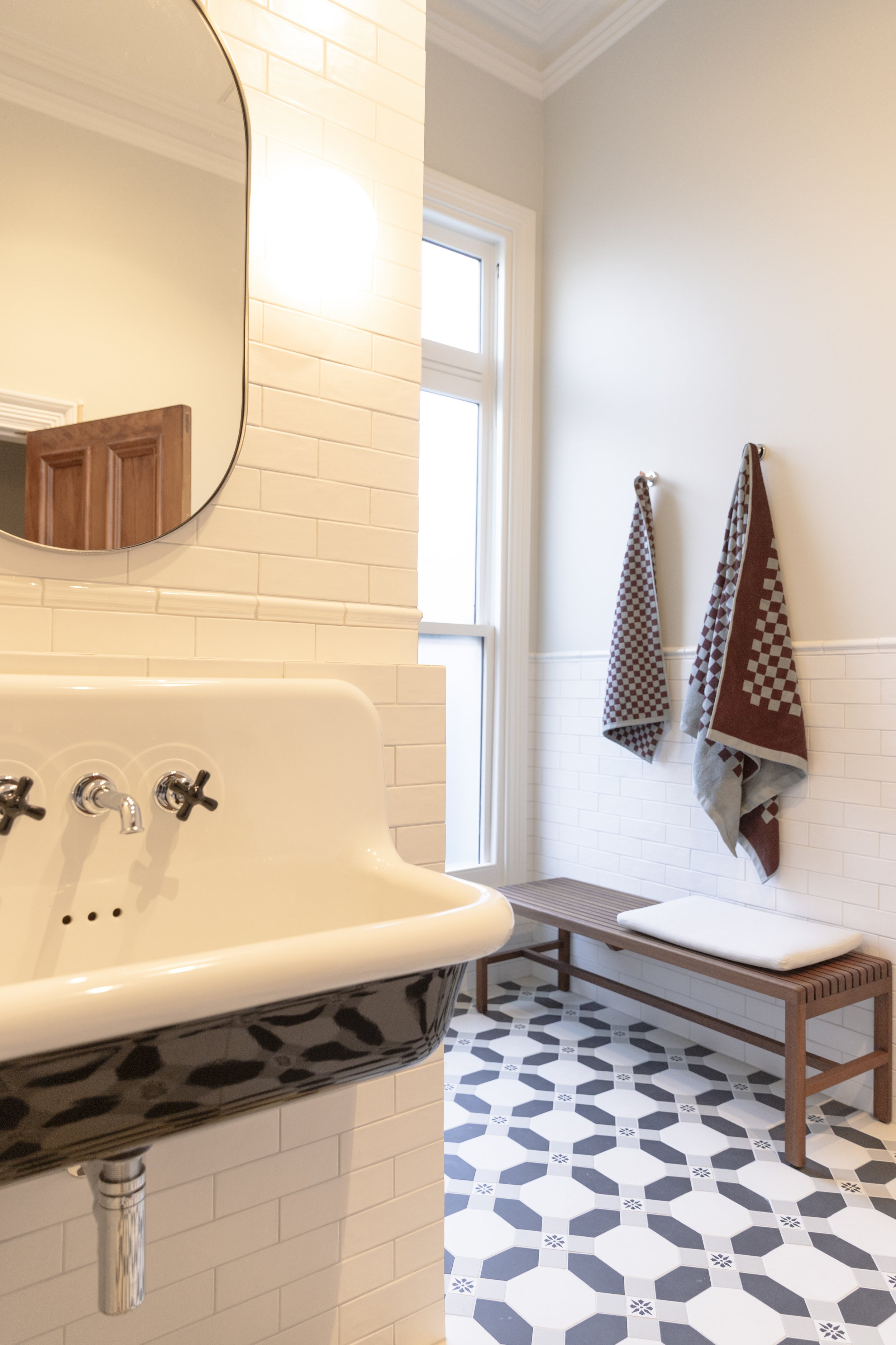 A cozy bathroom with a vintage white sink, black patterned exterior, and a large mirror. The room features white subway tile walls, a window with natural light, and a black and white hexagonal patterned floor. Two checkered towels hang on hooks on the wall, with a wooden bench below.