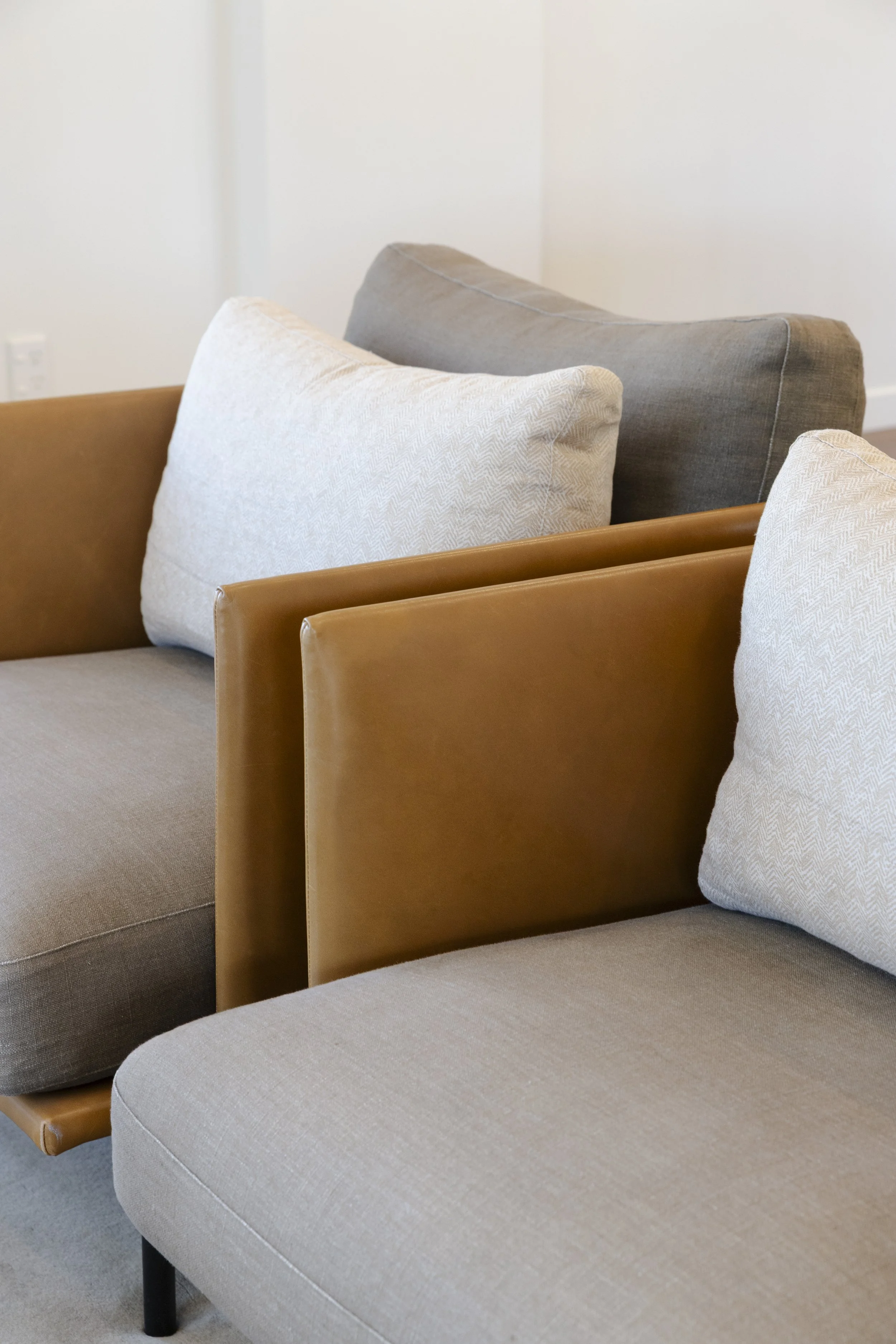 Close-up of beige, gray, and brown cushioned chairs with wooden armrests and fabric upholstery, arranged in a room with white walls.