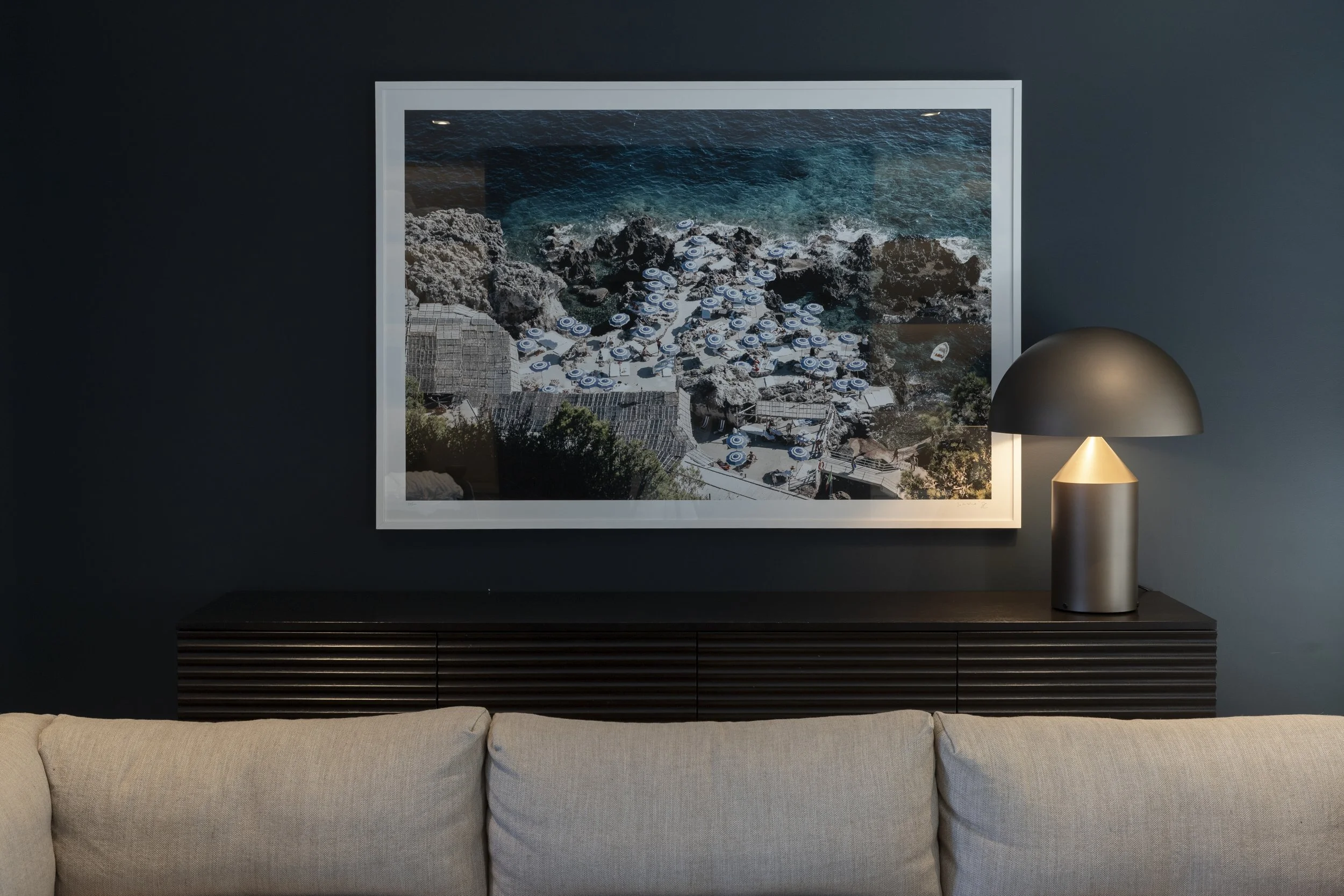Photograph of a framed picture of a rocky shoreline with blue umbrellas and a building on the coast, placed on a dark wall beside a modern table lamp, in a living room with a beige sofa.