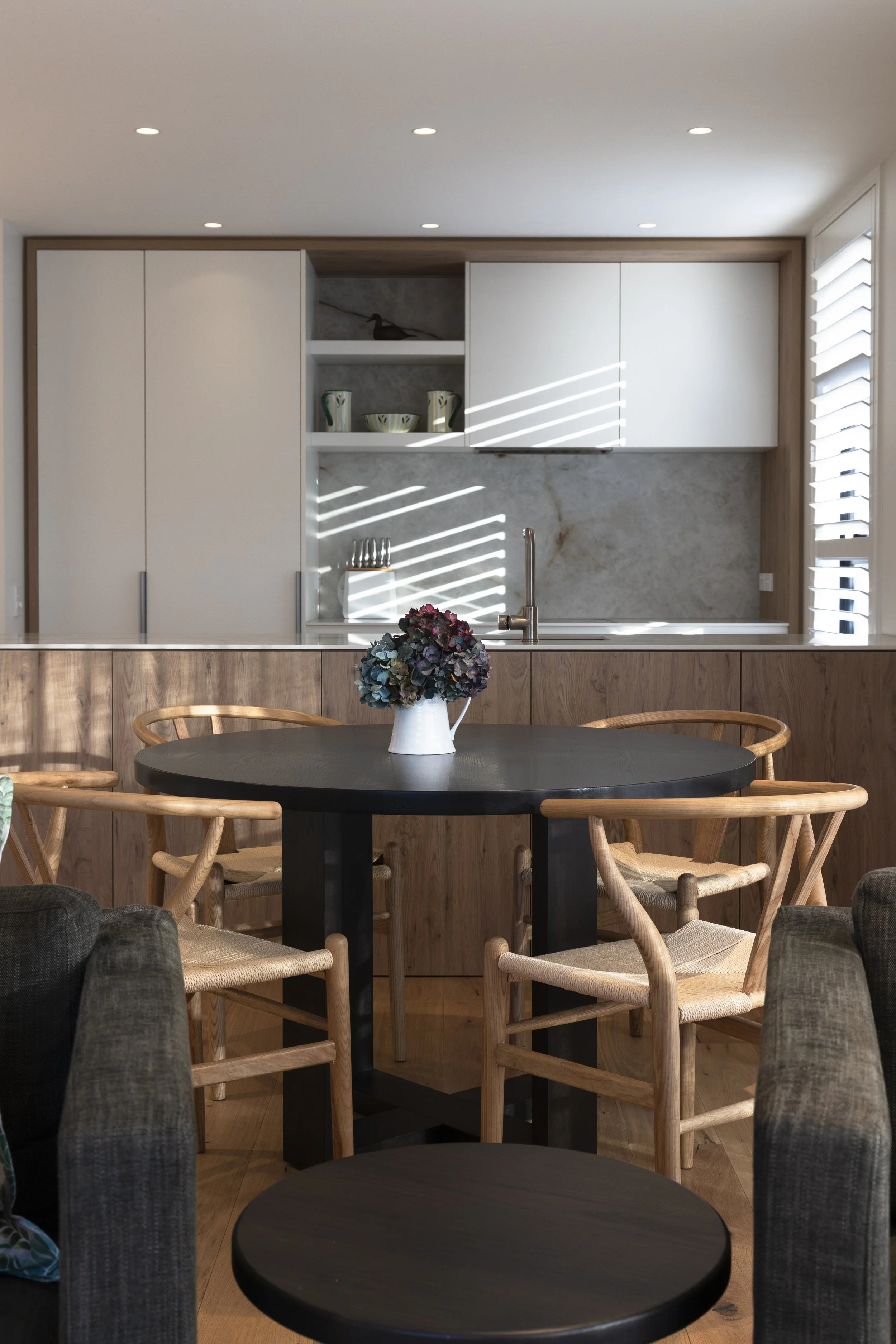 View of a modern kitchen with a black round table, floral centerpiece, wood chairs, white cabinets, open shelves with pottery, and natural sunlight through window blinds.