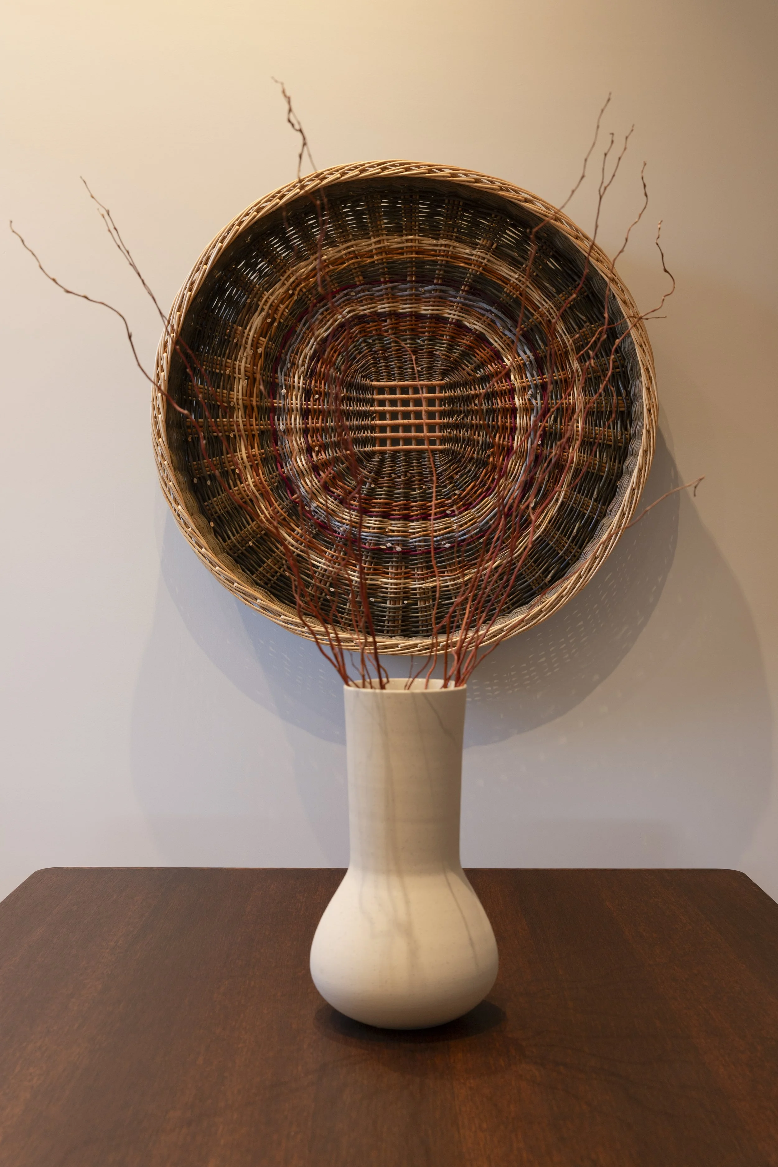 A woven basket with a white wooden base holding thin, curved, reddish-brown twigs, inside a white vase with a rounded bottom, on a dark wooden table against a plain beige wall.