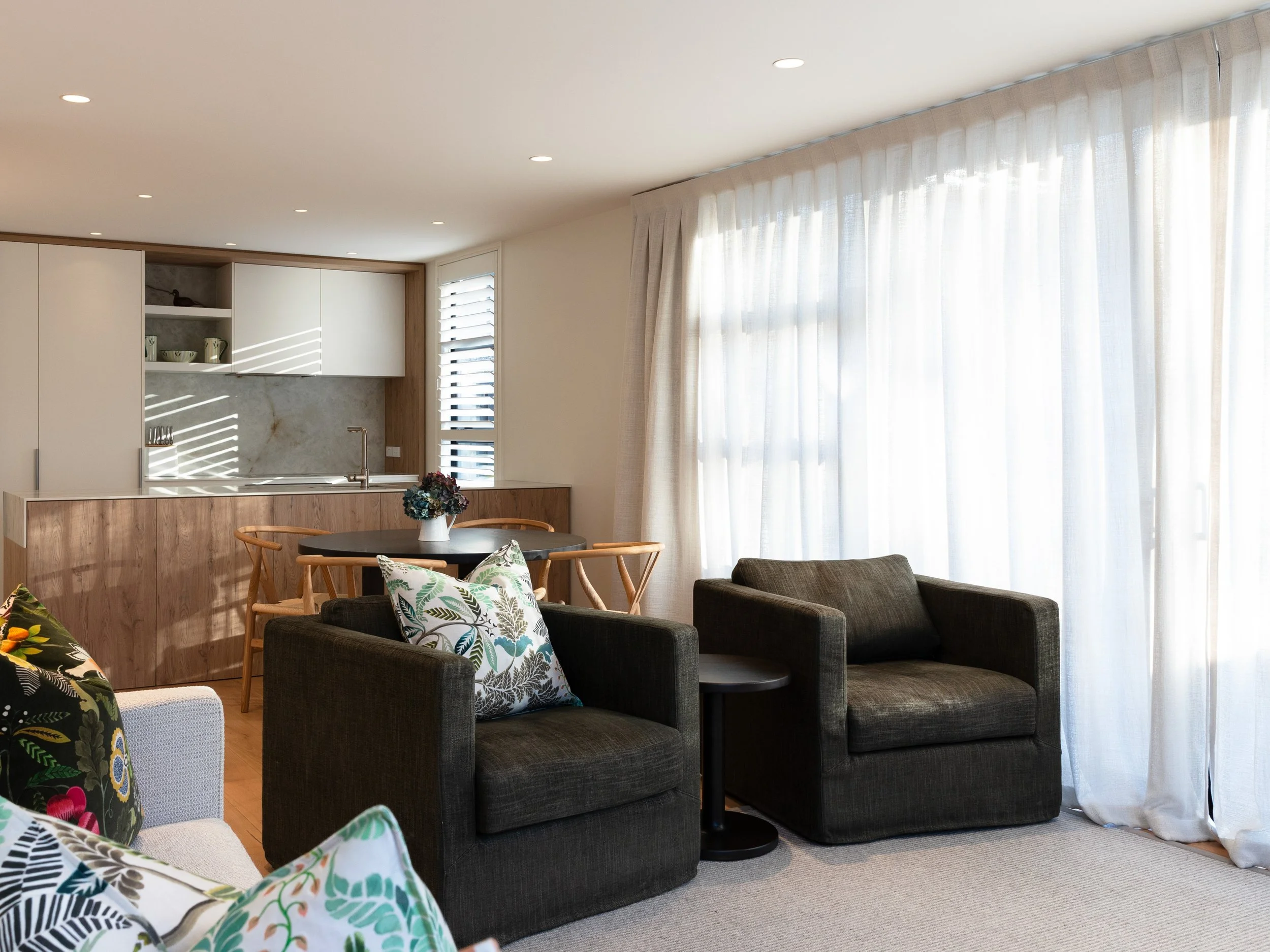 Living room area with dark gray sofas, floral and leaf-patterned throw pillows, a small round black table, a round dining table with wooden chairs, a vase with flowers, white curtains over large windows, white kitchen cabinets with open shelves, and a marble backsplash.