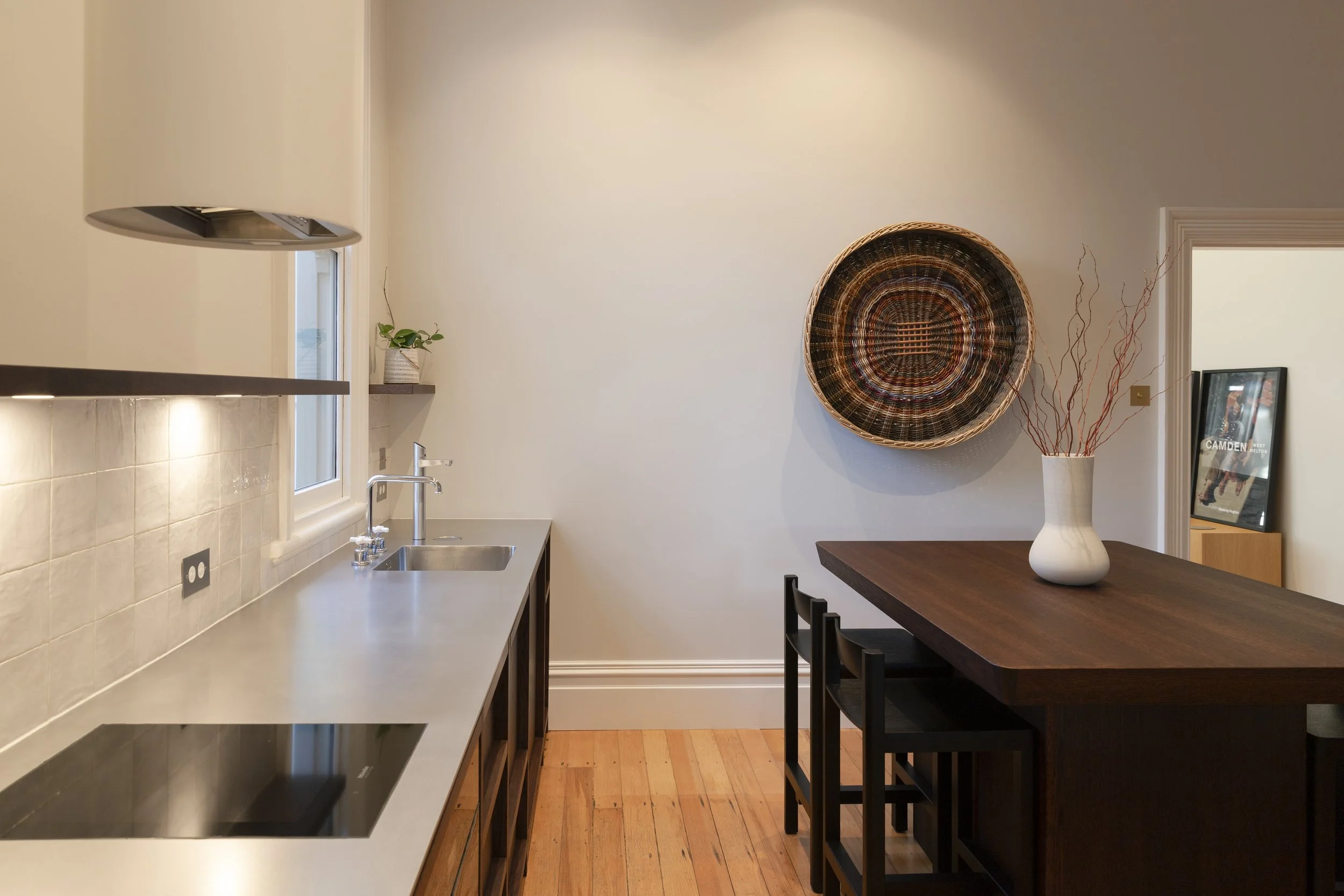 A kitchen with a white quartz countertop, a stainless steel sink, and beige tiled backsplash. There is a small window above the sink, a small plant on a shelf, and a black induction cooktop on the counter. A dark wooden dining table with black chairs is adjacent, with a large woven basket on the wall and a white vase with decorative branches on the table.