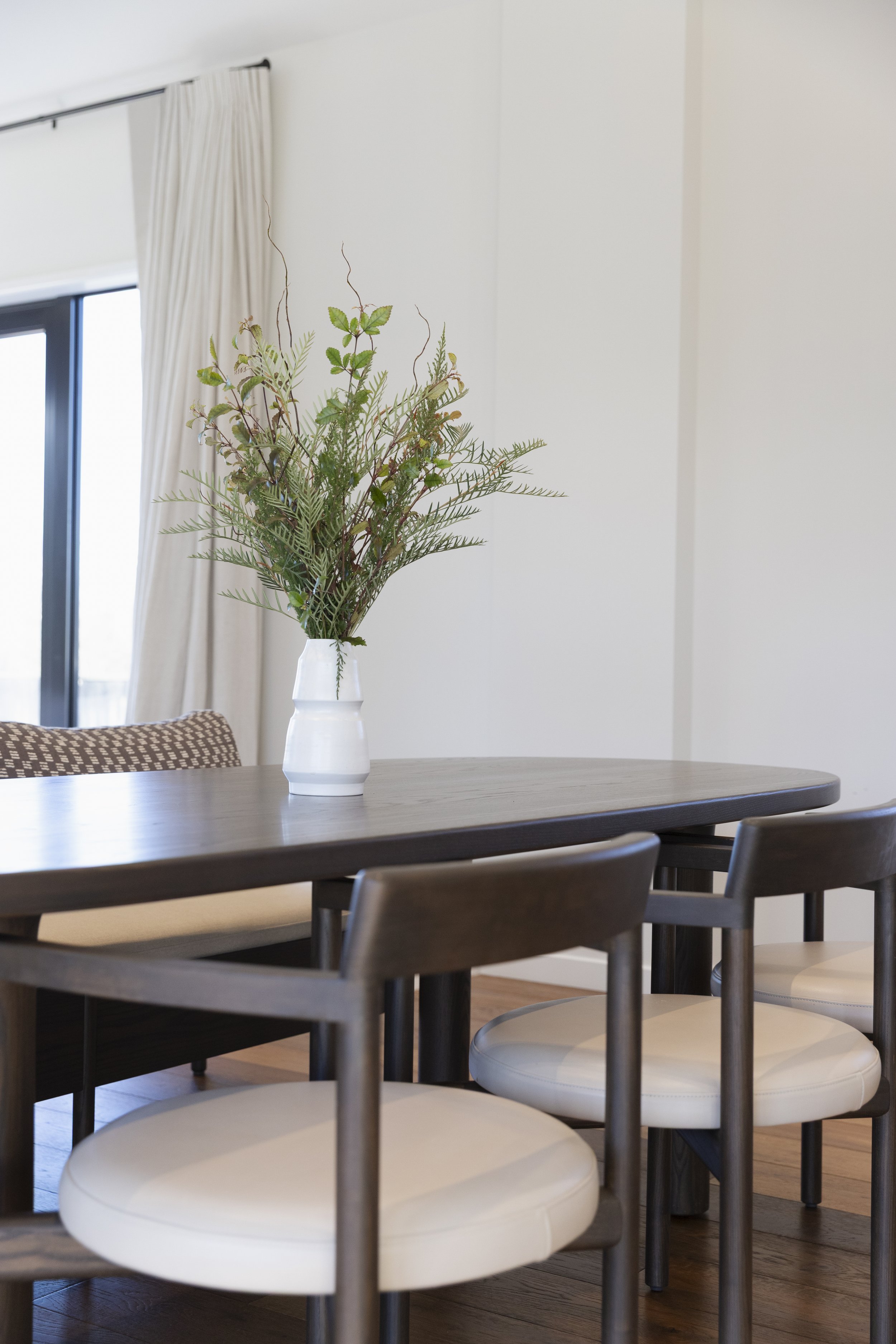 A dining room table with dark wood finish, surrounded by chairs with grayish frames and white cushioned seats. A tall white vase with green foliage is on the table. There is a wall with beige curtains covering a window or sliding door in the background.