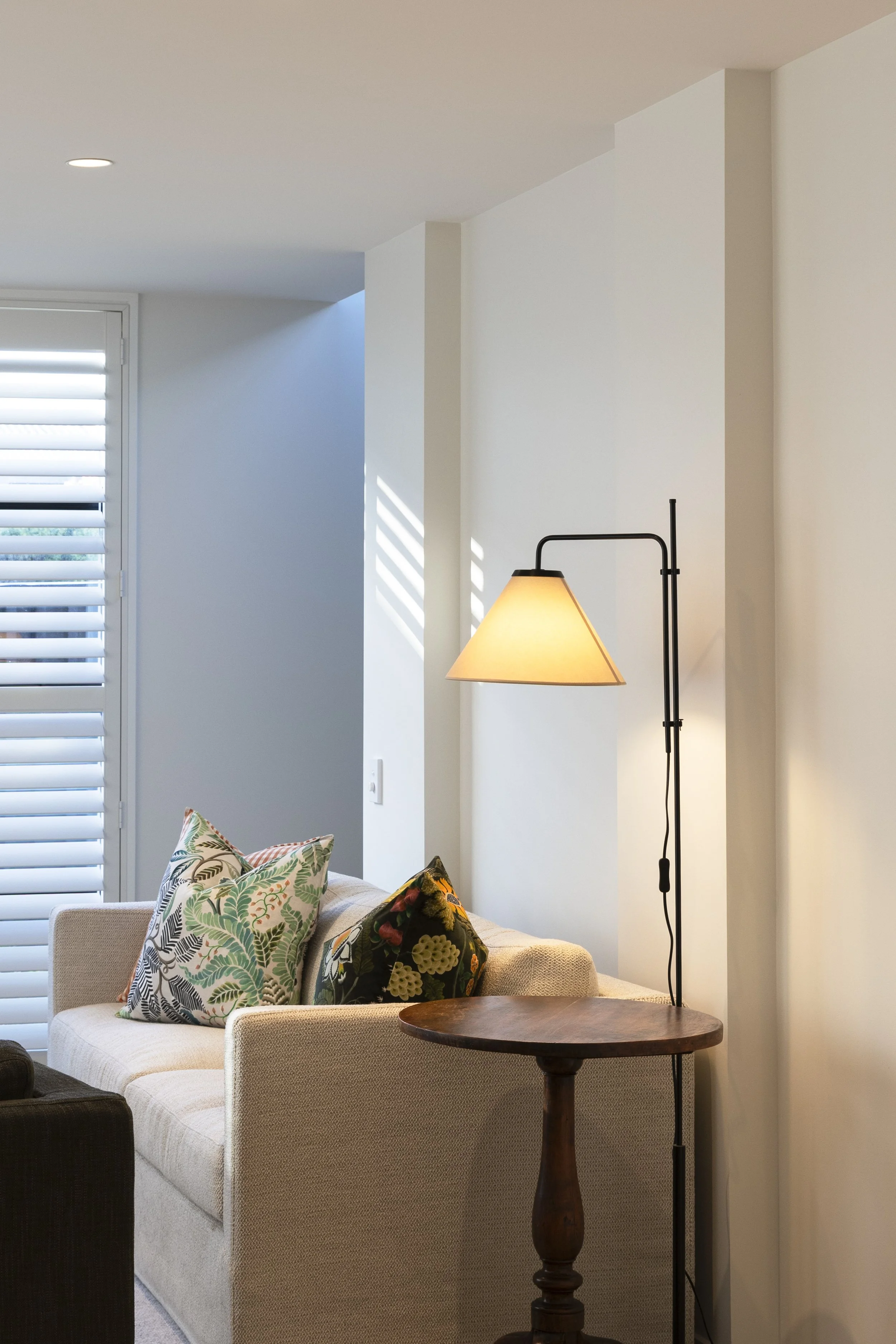 Living room with beige sofa, patterned pillows, wooden side table, and floor lamp with beige shade, near a window with white blinds, and a white wall with ceiling lighting.