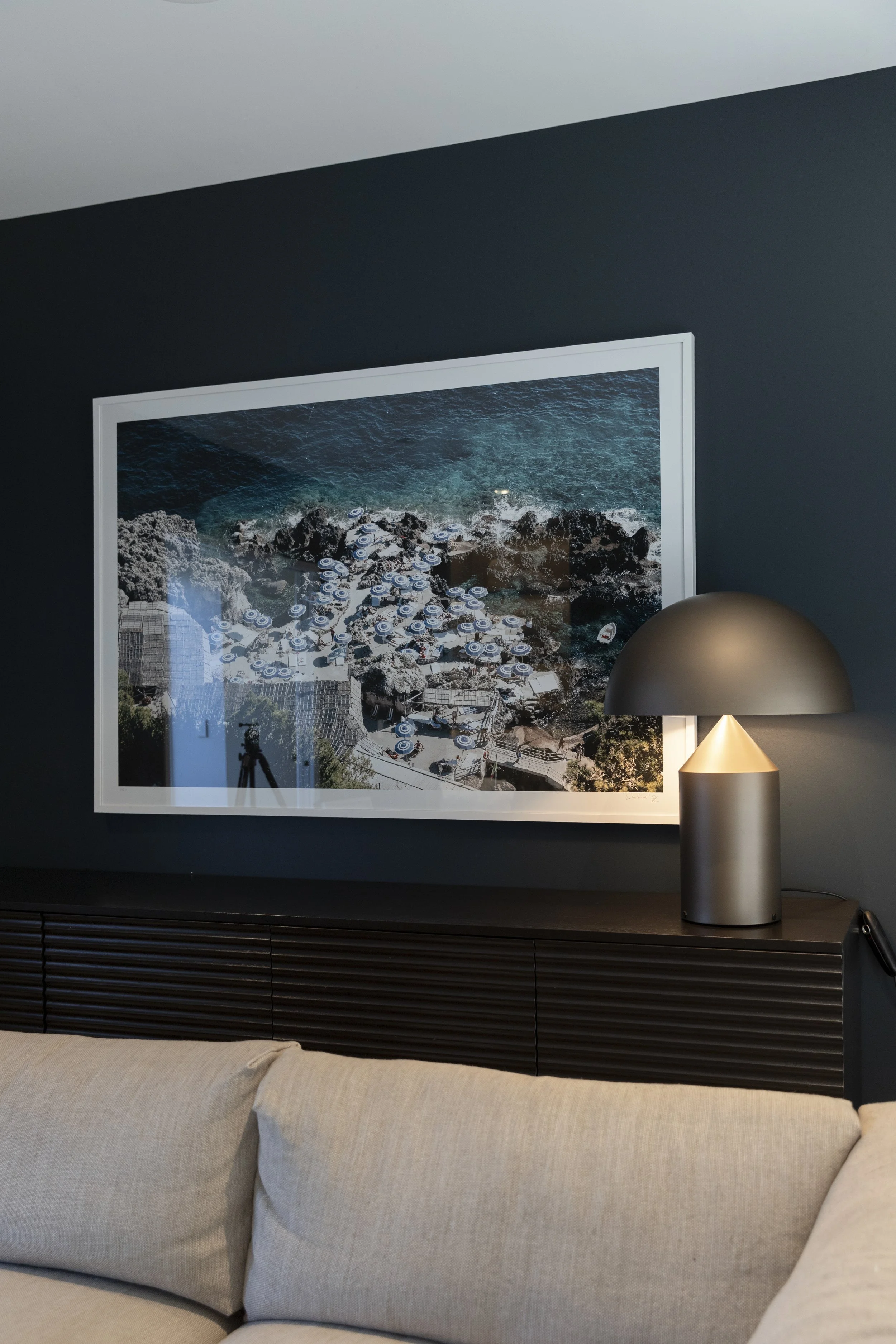 A framed aerial photograph of a rocky coastline with water and umbrellas, hanging on a dark wall in a living room, with a modern lamp and beige sofa in the foreground.
