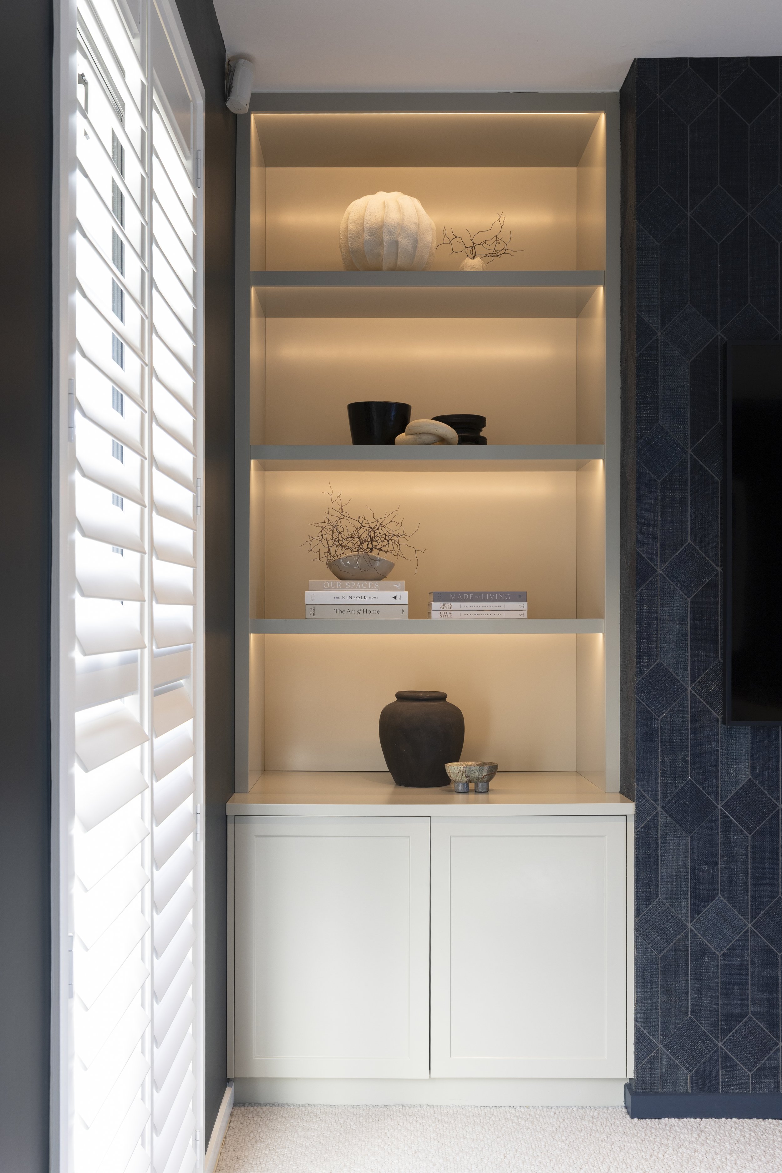 Built-in display cabinet with decorative objects including pottery and books, next to a window with plantation shutters and a textured dark accent wall.