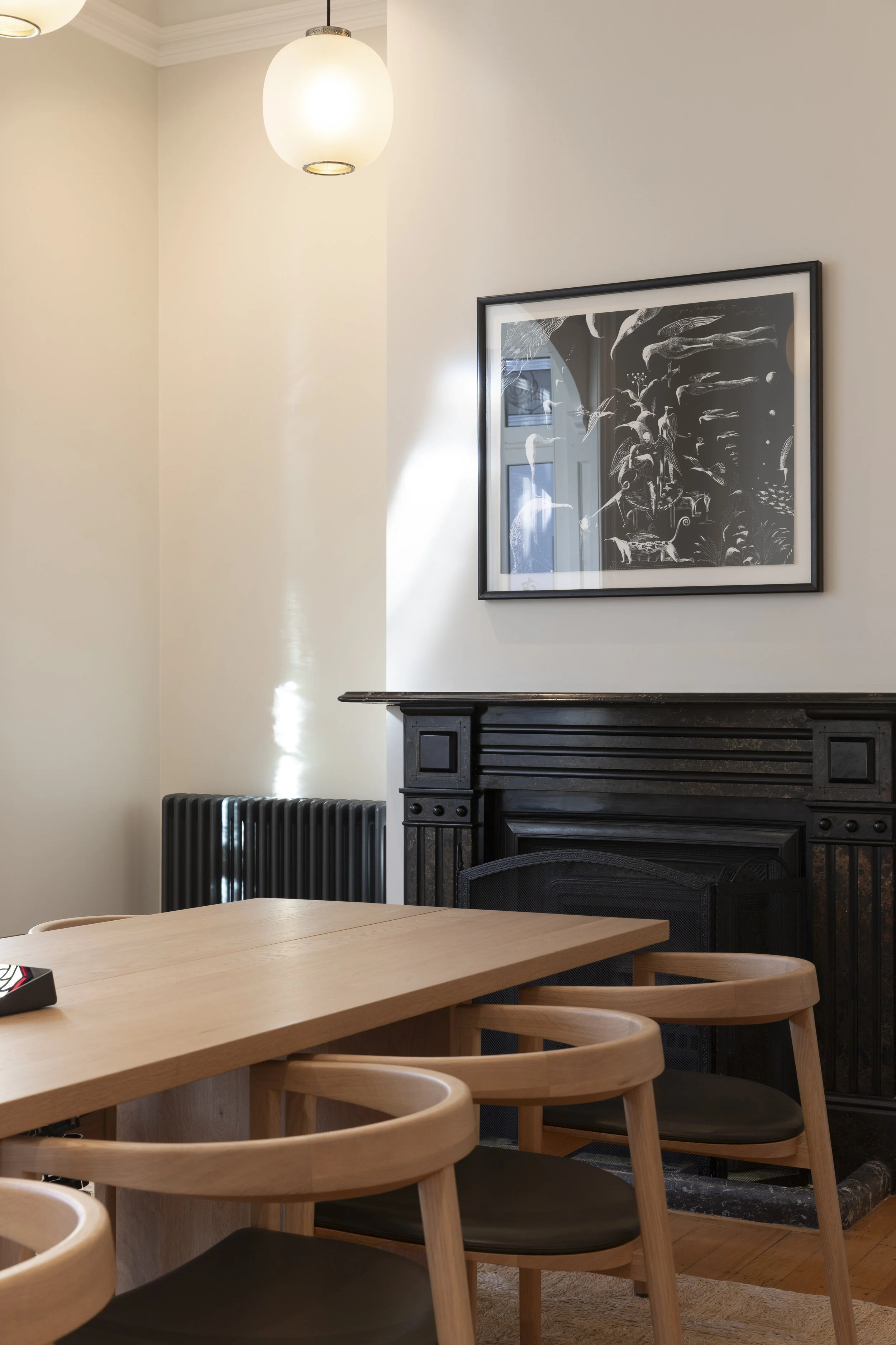 Interior of a dining room with a light wooden table surrounded by matching chairs. A black fireplace with decorative carvings is against the wall, above which hangs a framed black-and-white art print. A white ceiling light fixture hangs from the ceiling. Part of a black radiator and a partial view of another room are visible.