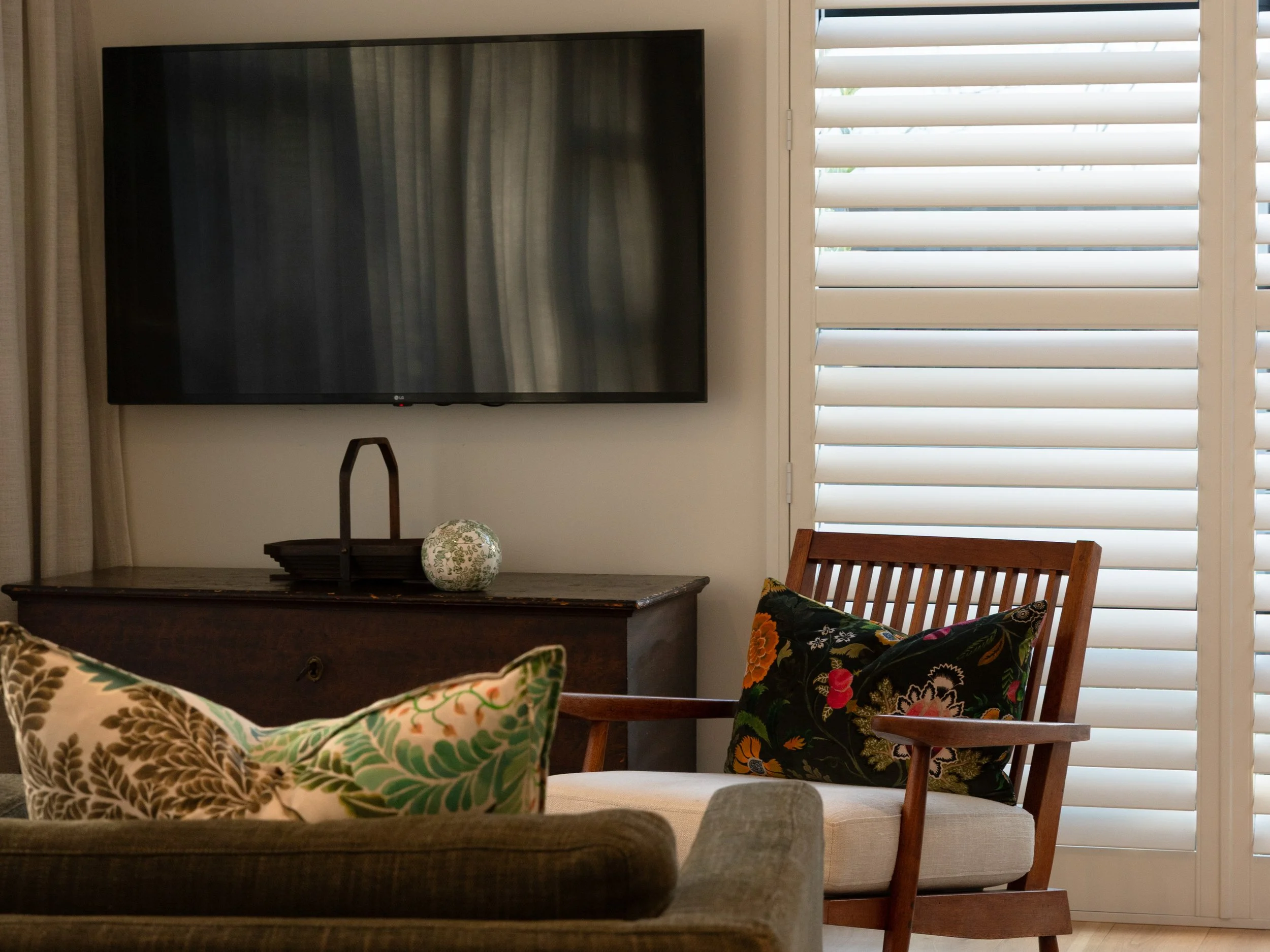 Living room with flat-screen TV, wooden console table, patterned armchair, and window with white blinds.