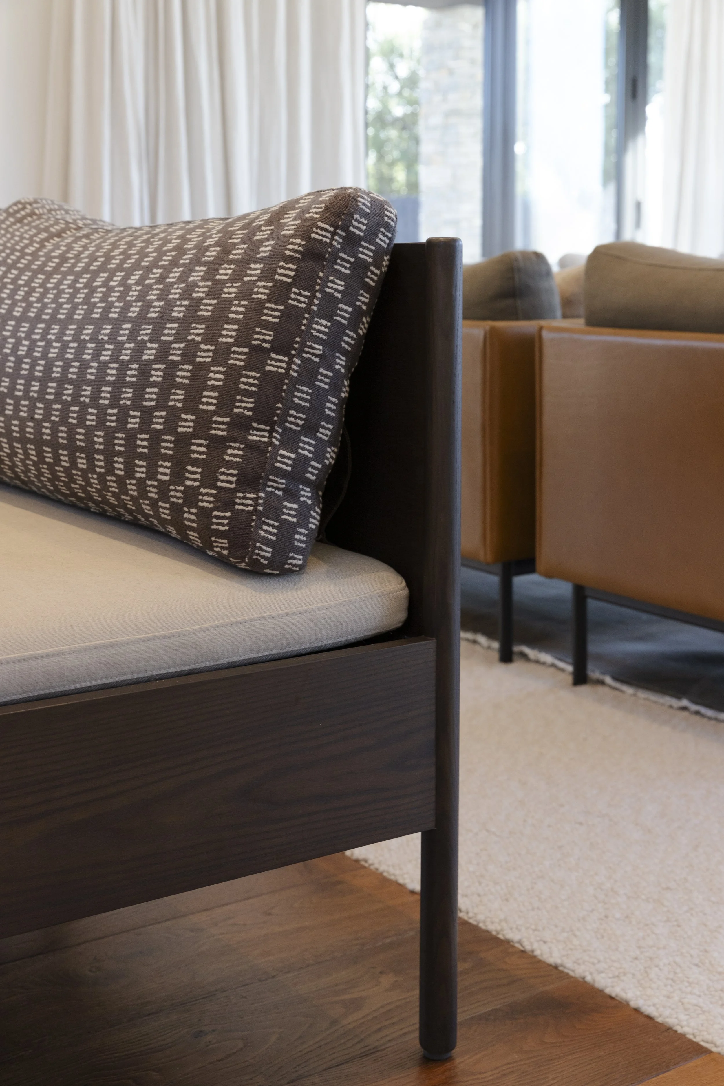 Close-up of a bed with a patterned pillow in a bedroom with large windows, curtains, and a view of trees outside.