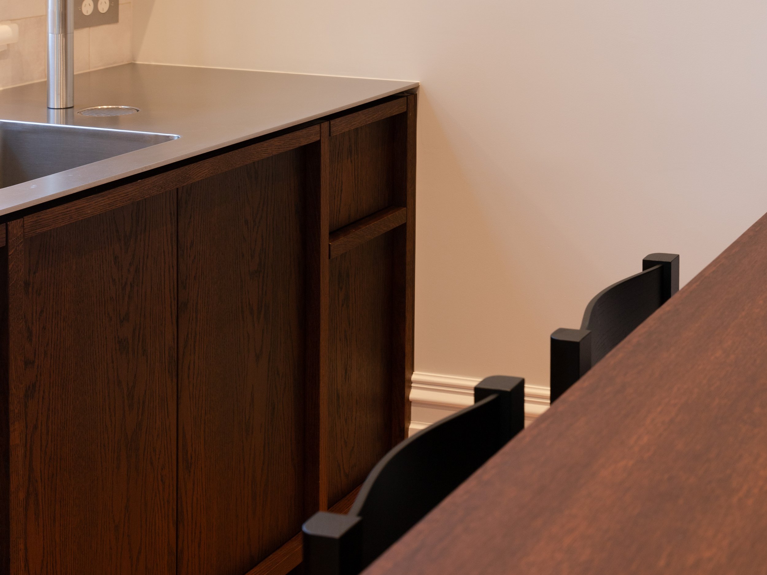 Part of a kitchen counter with a dark wood cabinet, a stainless steel sink, and a beige wall with electrical outlets. Part of a chair is visible in the foreground.