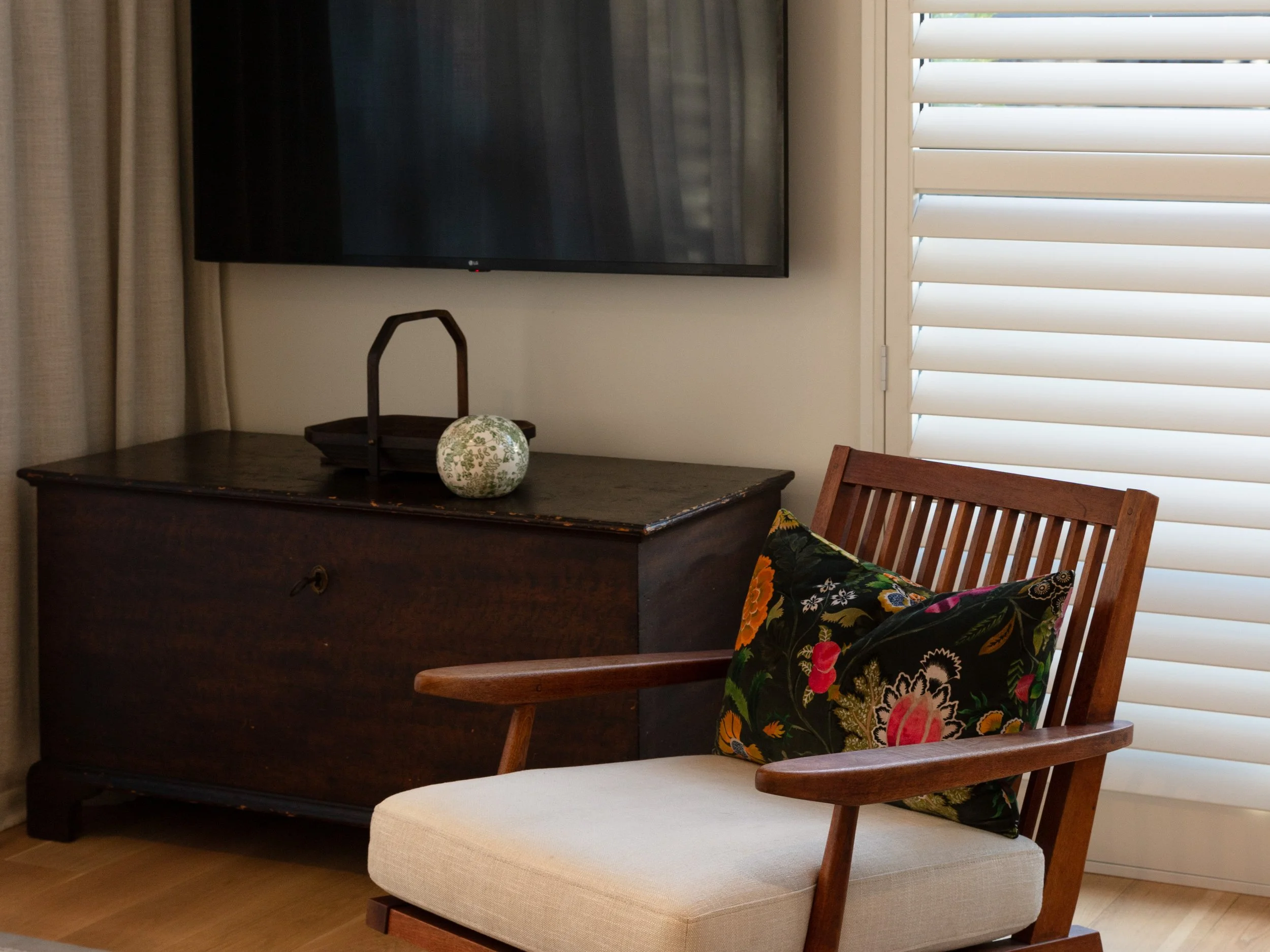 Living room corner with a dark wooden dresser, wall-mounted flat-screen TV, a wooden chair with floral cushion, beige ottoman, and window with white plantation shutters