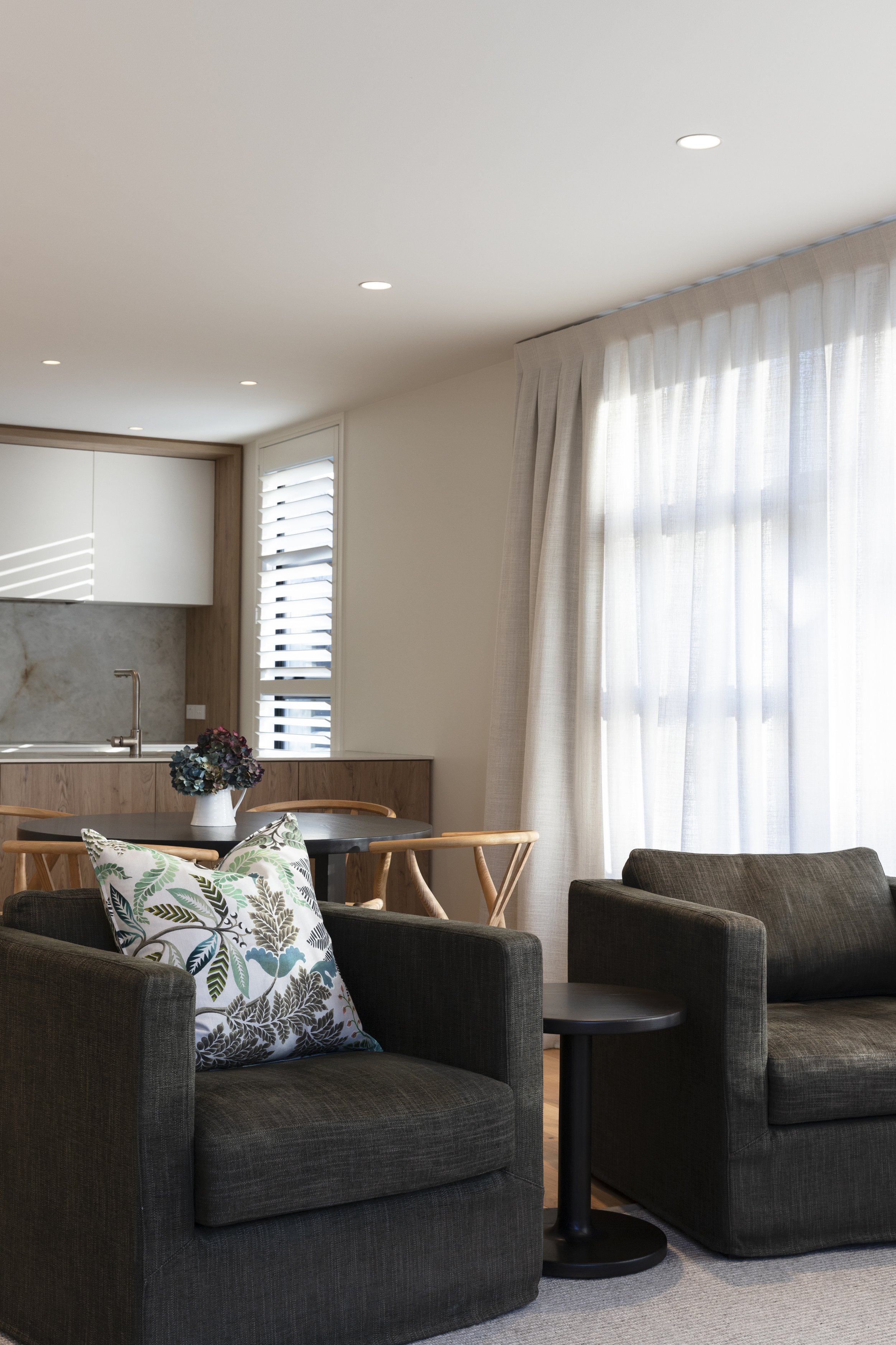 A modern living room area featuring two dark upholstered armchairs and a dark round side table. There is a patterned pillow with a botanical design on one armchair. The background includes a dining table with wooden chairs, a window with white blinds, sheer curtains, and a kitchen area with white cabinetry, a marble backsplash, and wooden accents. The ceiling has recessed lighting, and natural light fills the room.
