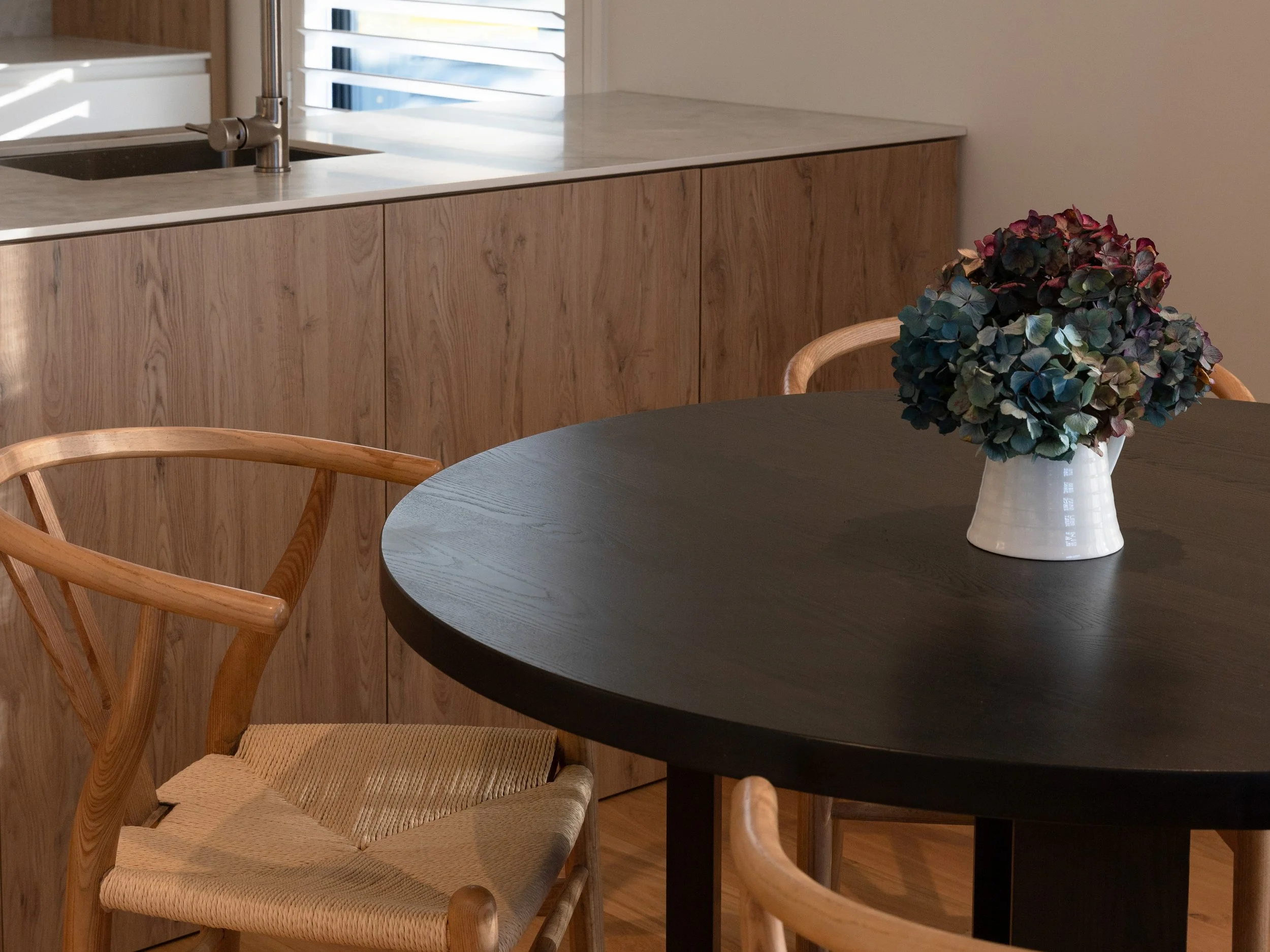 A round black dining table with a vase of multicolored hydrangeas, surrounded by wooden chairs, in a room with wooden cabinetry and a window with blinds.