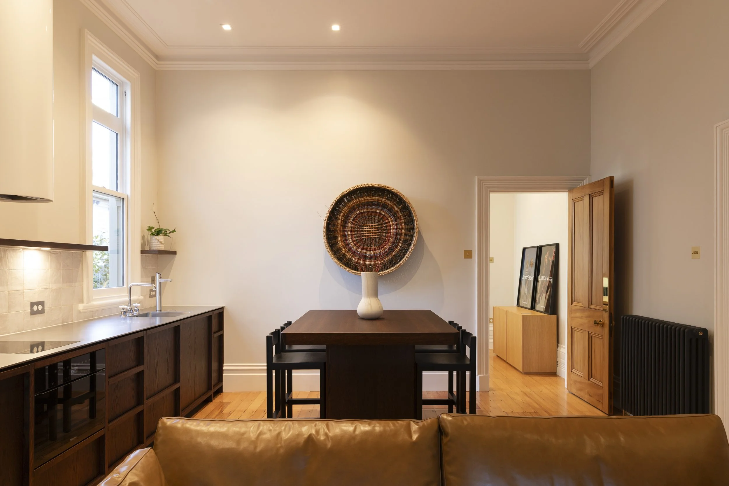 Interior view of a modern dining room with a wooden table, black chairs, a large round woven wall decor, a white vase, a window with white trim, and a doorway leading to another room. There is also a kitchen counter with a sink and potted plant.