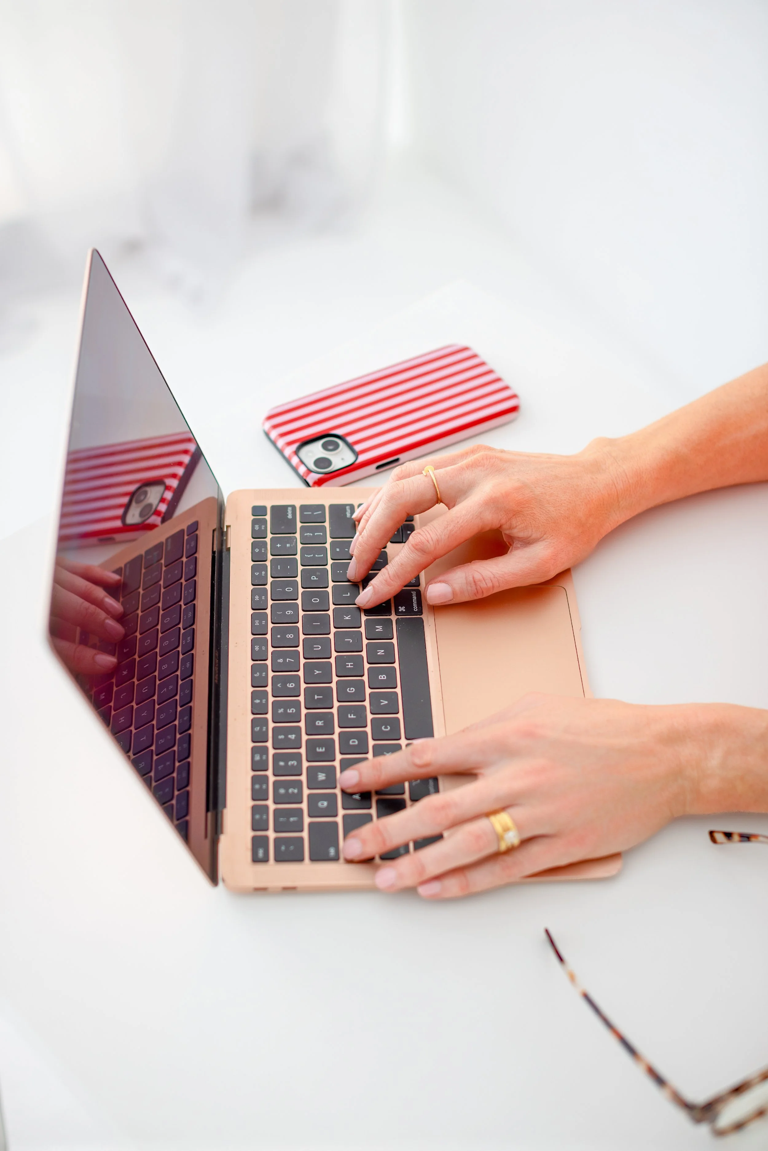 Person using a laptop with hands on the keyboard, a striped red and white phone case on the desk, white surface, and glasses partially visible in the corner.